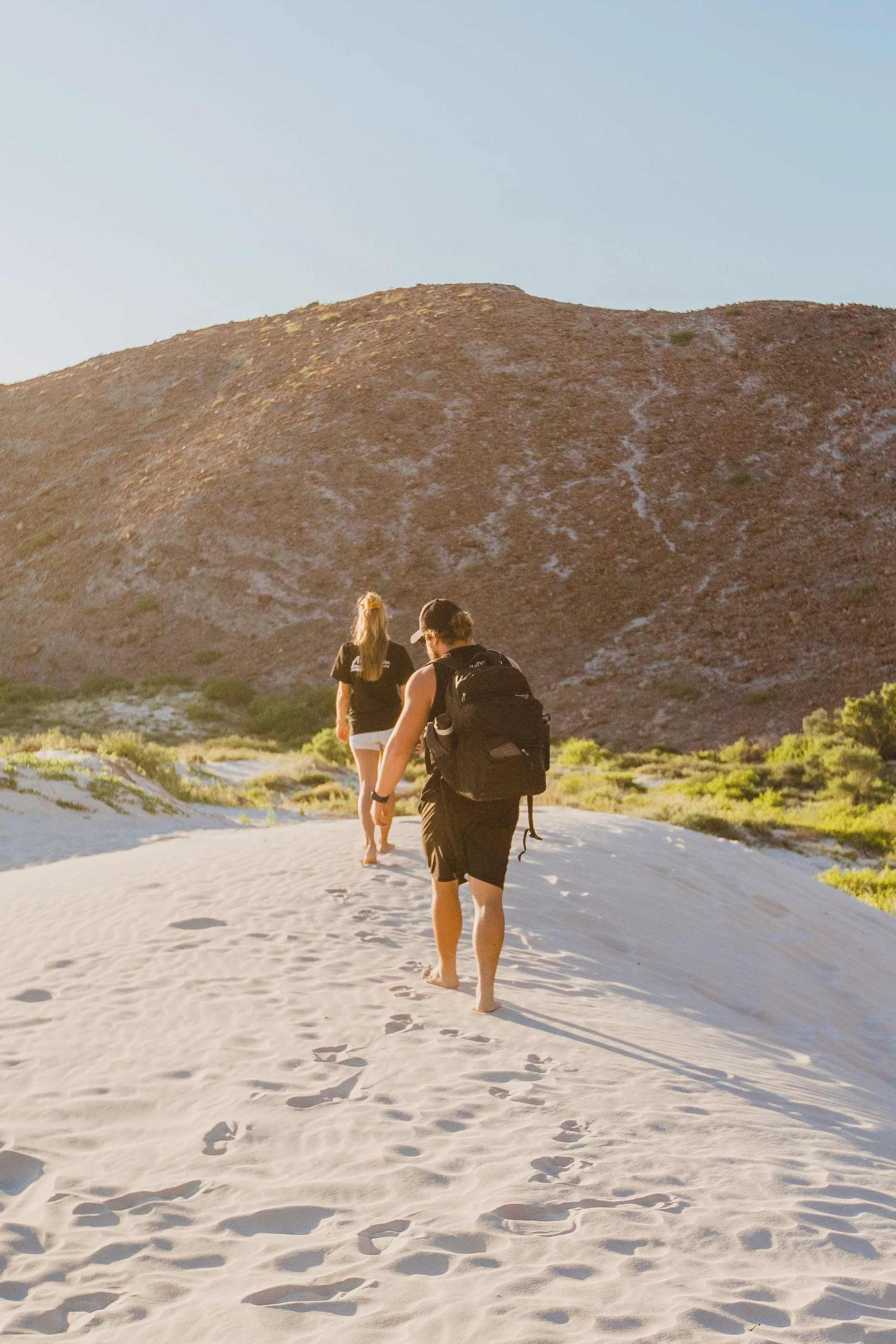 Two people walking on a sandy trail in a desert landscape with low green vegetation, carrying backpacks, with a large mountain in the background under a clear sky.
