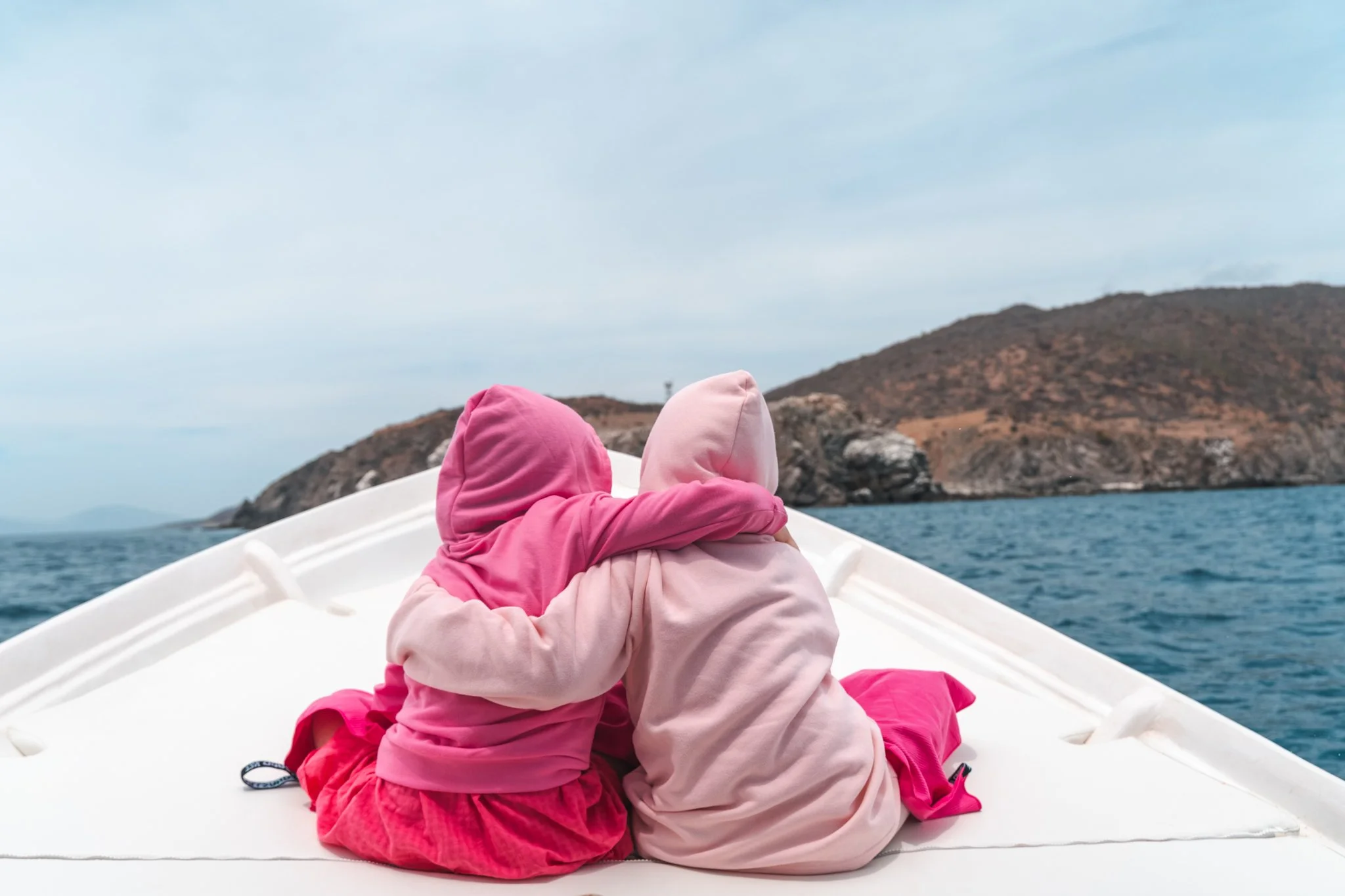 Two children wearing pink and beige hooded jackets sitting close together on a boat, with their arms around each other, looking at the water and distant rocky hills.