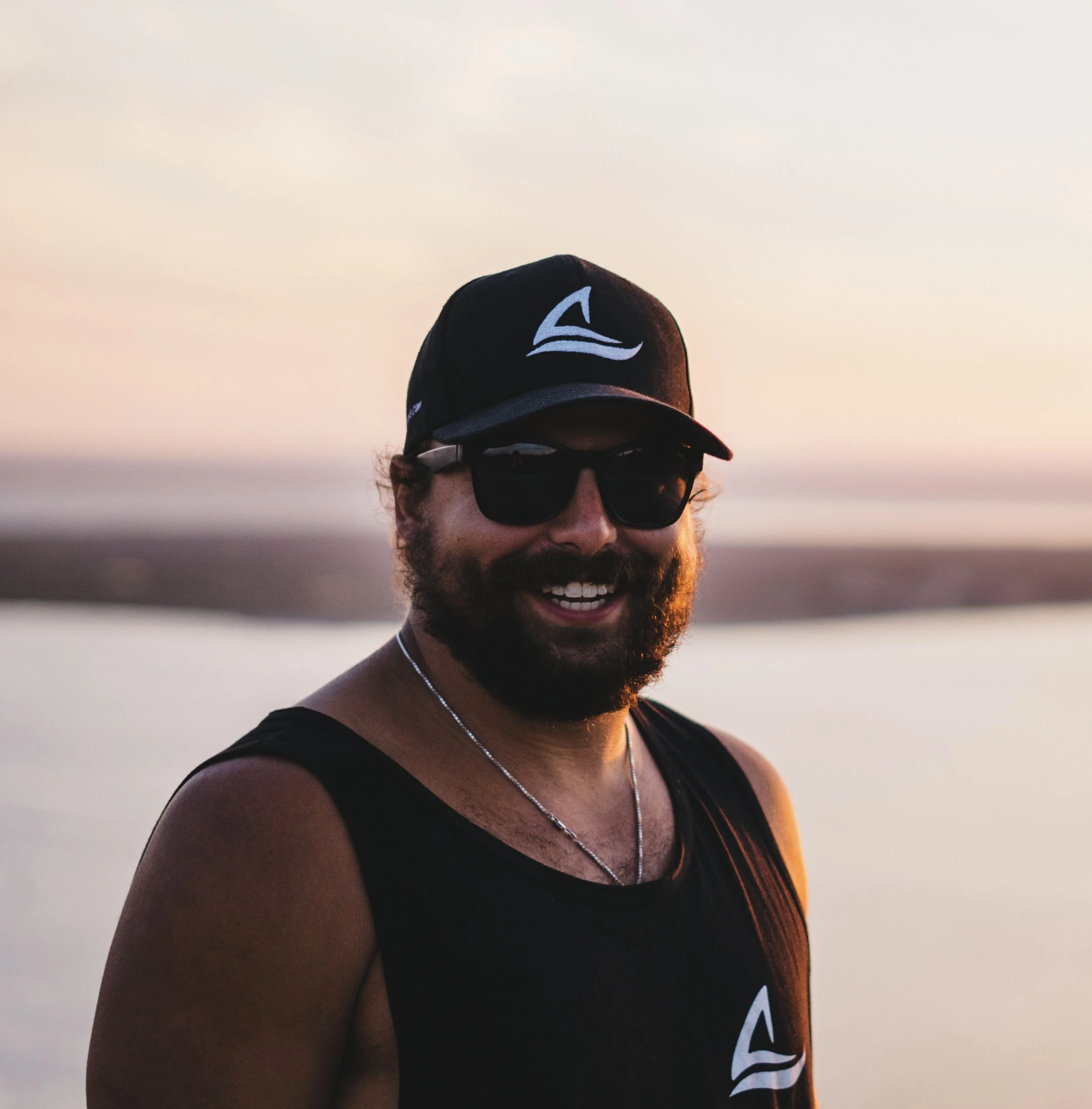 Man smiling on a beach at sunset, wearing sunglasses, a black cap with a logo, a black tank top, and a chain necklace.