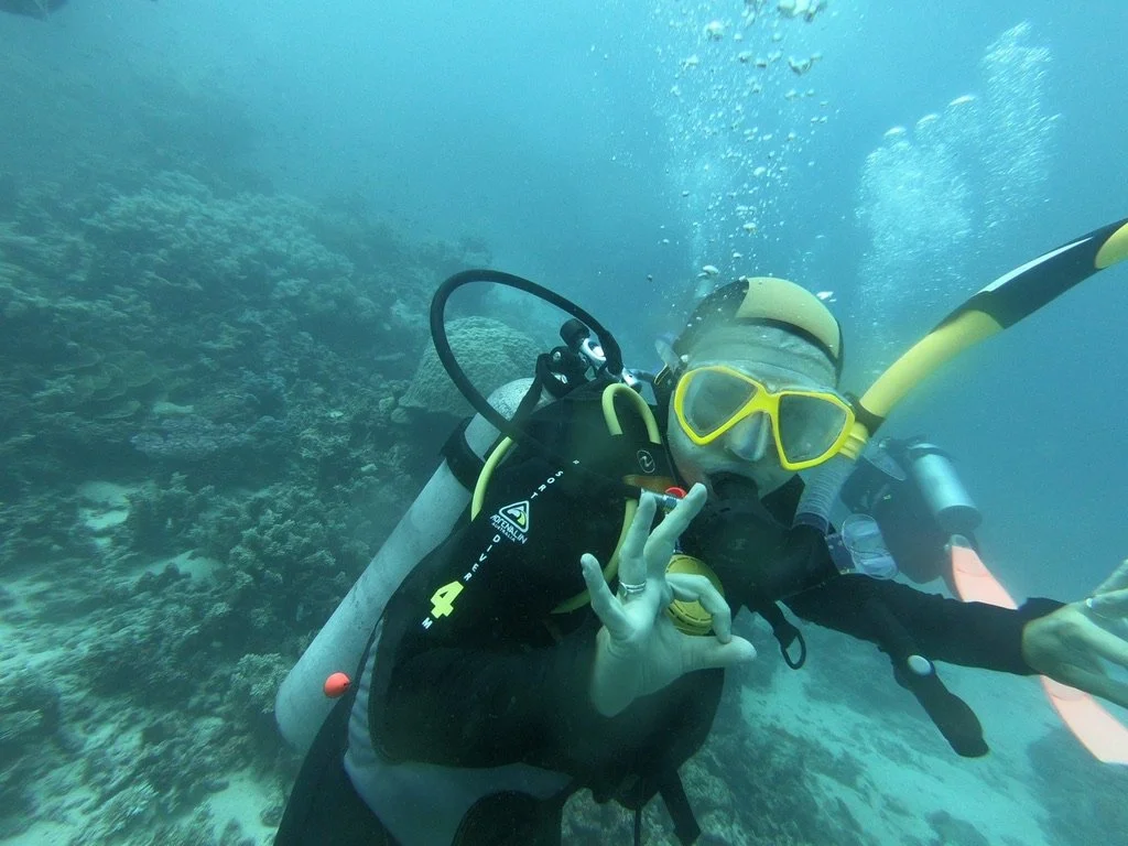 A person scuba diving underwater, making an OK hand signal, wearing a yellow mask and snorkel, with coral visible in the background.