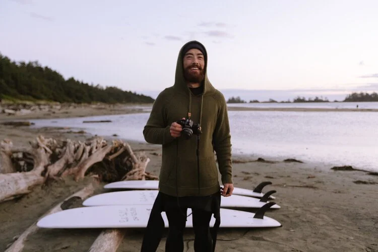 Smiling man in a green hoodie holding a camera standing on a beach with five paddleboards lined up on the sand behind him, near a body of water with trees in the background.