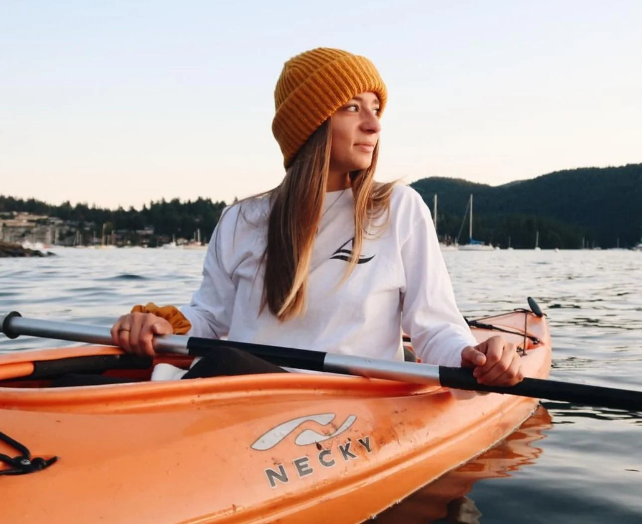 Young woman kayaking on a calm body of water during sunset, wearing a yellow knit beanie and a white long sleeve shirt with a black logo.