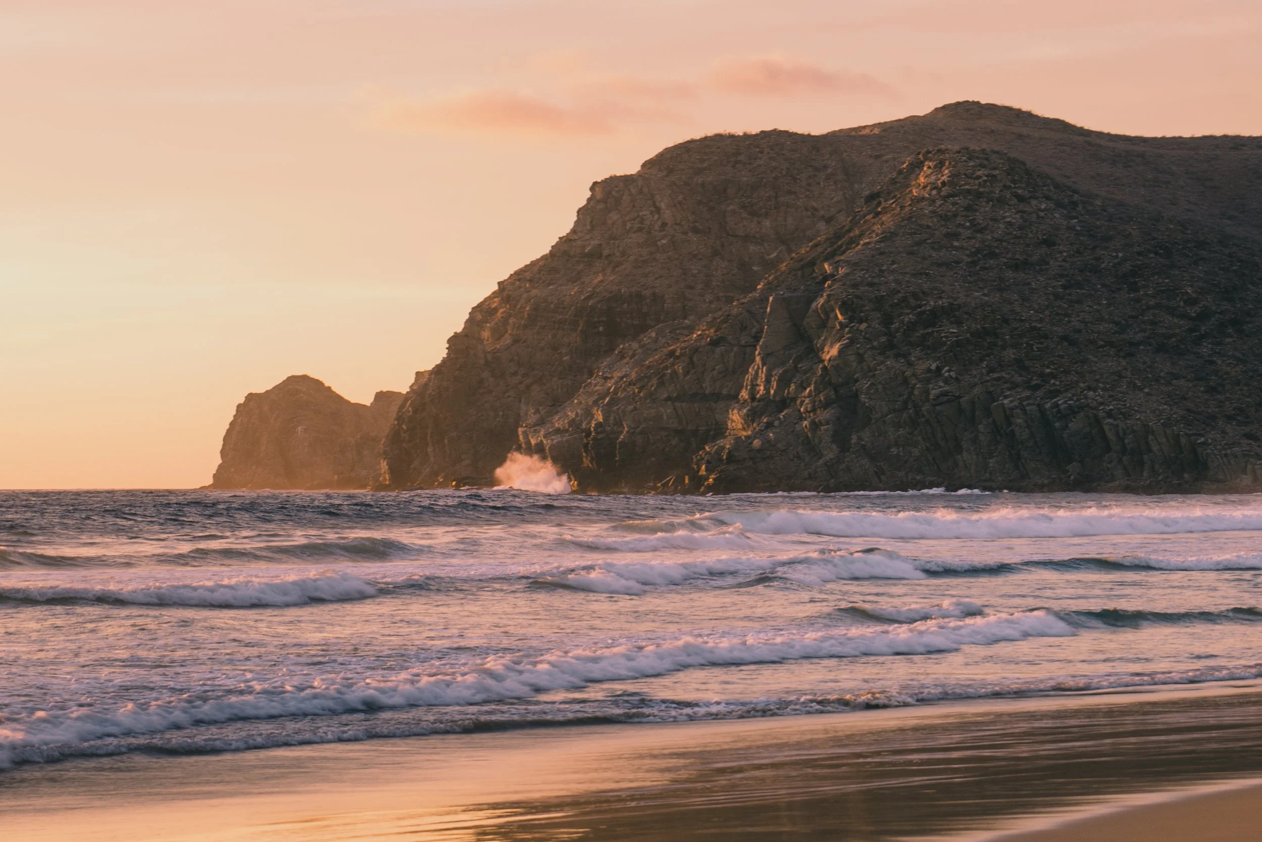 Sunset over the ocean with waves hitting the shore and rocky cliffs in the background.