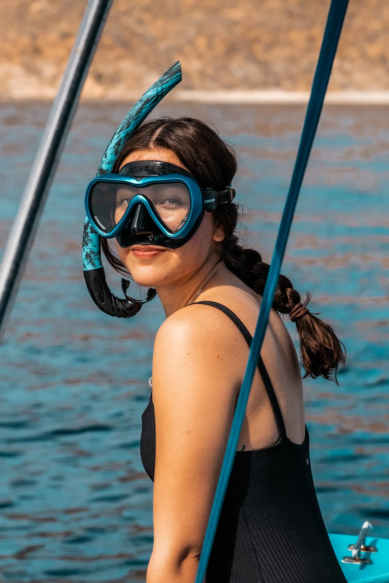 A woman wearing a snorkeling mask and fins on a boat, looking over her shoulder at the camera, with water and a rocky shoreline in the background.