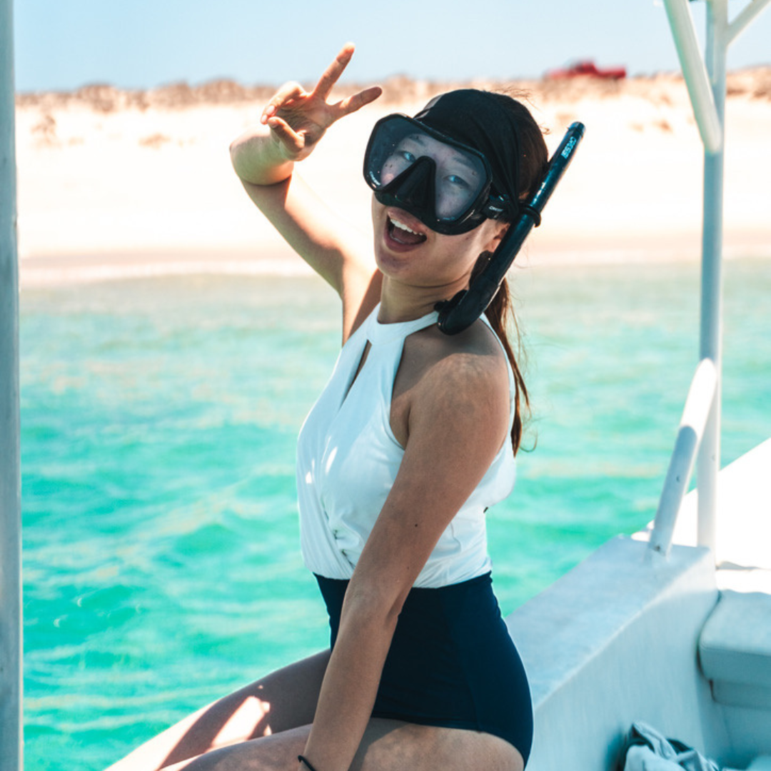 Woman in a white top and black swimsuit with a snorkel and mask smiling and making a peace sign while sitting on a boat, with clear turquoise water and sandy beach in the background.