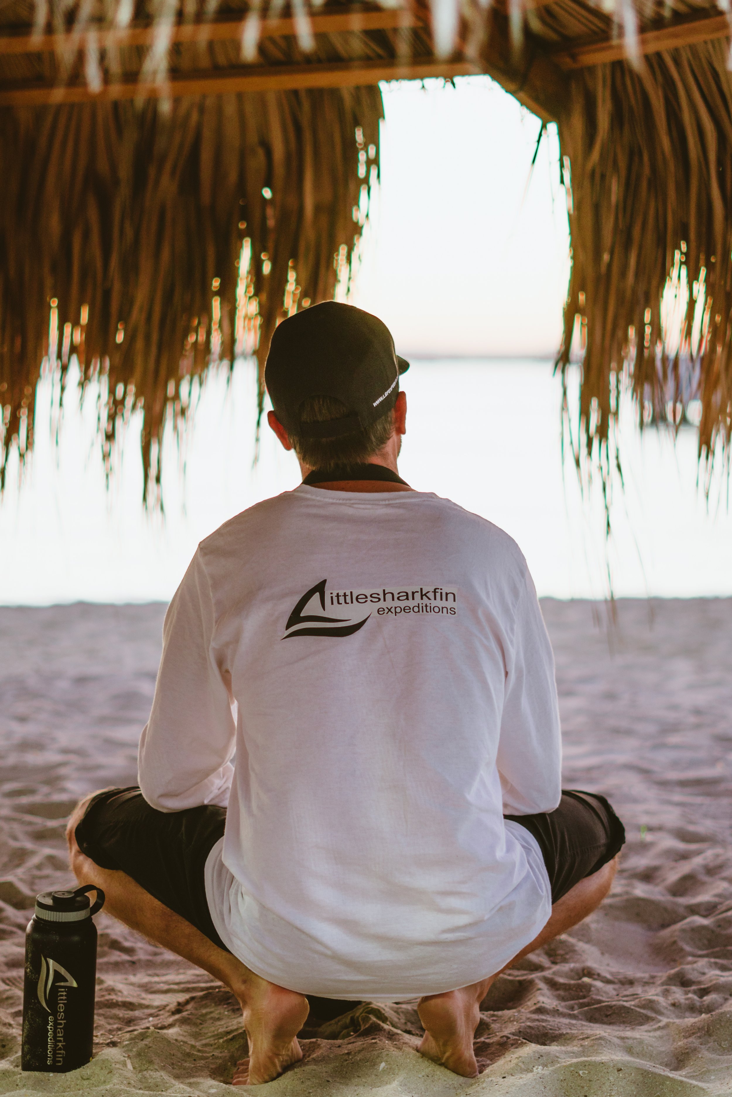 A man sitting cross-legged under a thatched hut on the beach, facing the ocean, wearing a white shirt with 'Littlesharkfin expeditions' logo, a black cap, and black shorts, with a black water bottle beside him.