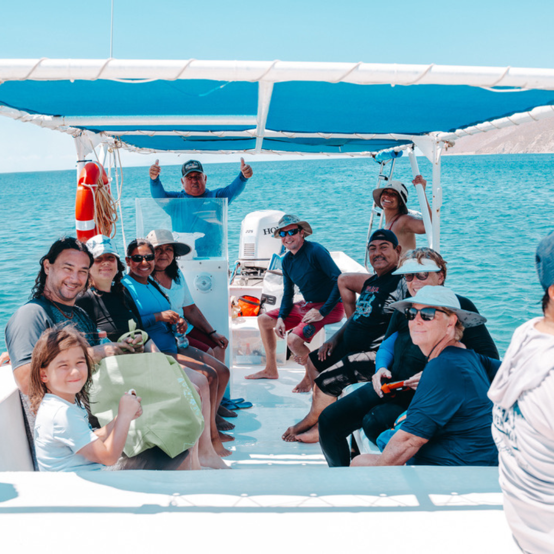 A group of people on a boat enjoying a sunny day, with some giving thumbs up and others smiling, surrounded by water and a distant shoreline.