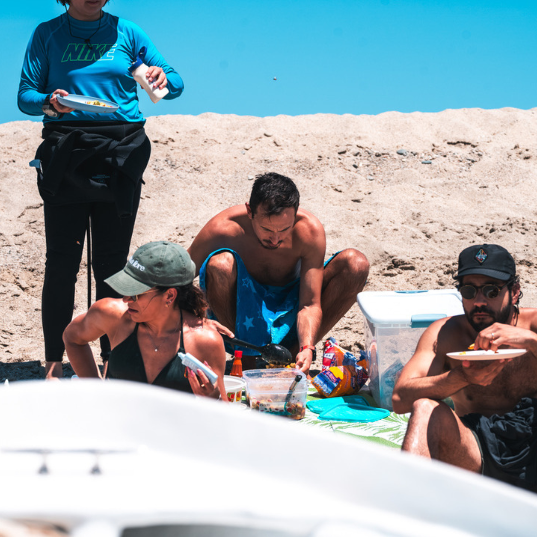 Group of four people on a sandy beach enjoying a meal together, with one person sitting in the background and three in the foreground, some wearing hats and sunglasses.