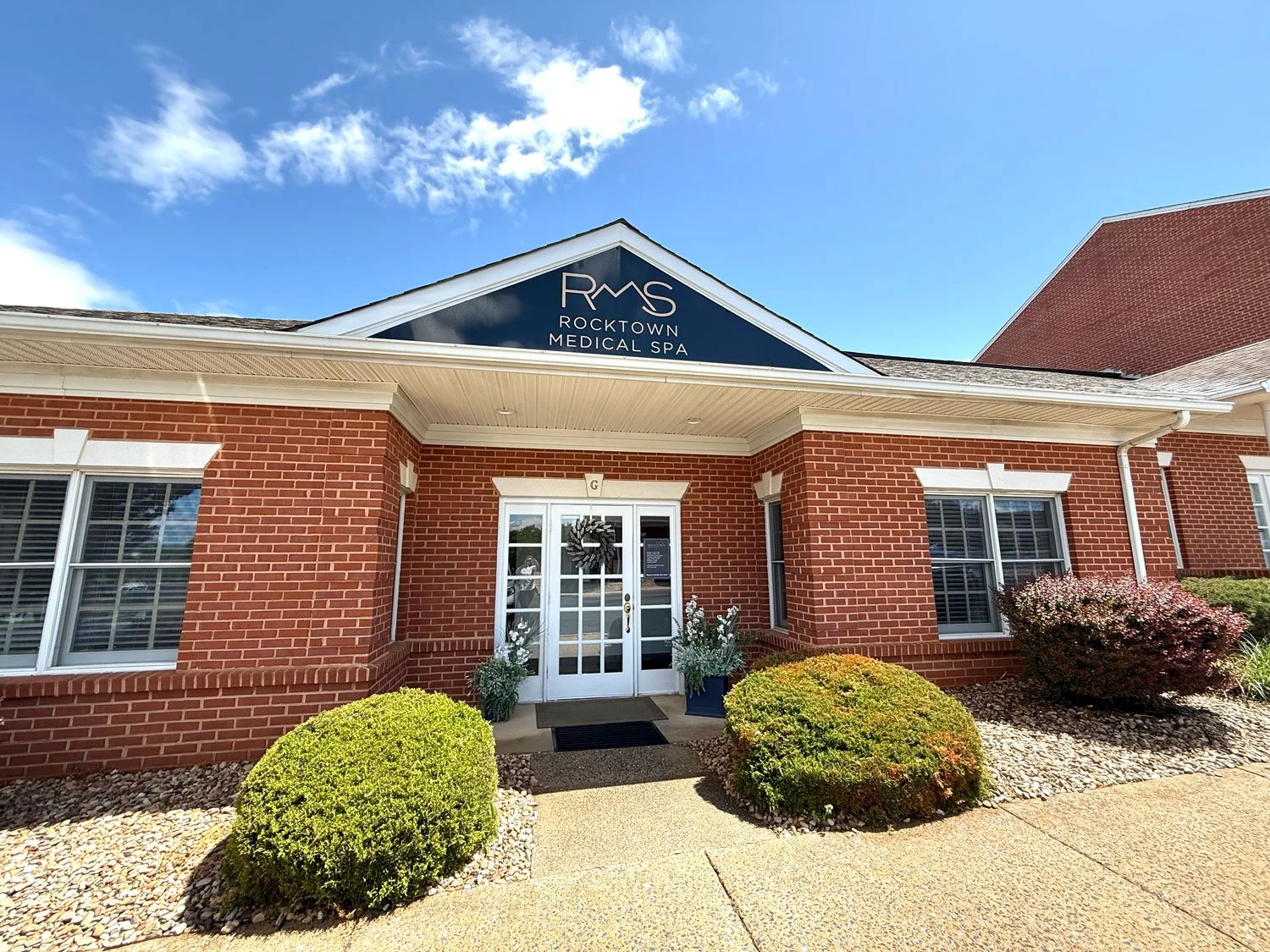 Red brick building with a blue triangular sign reading "RMS Rocktown Medical Spa" above the entrance, two white doors with a wreath, and well-manicured bushes outside on a sunny day.