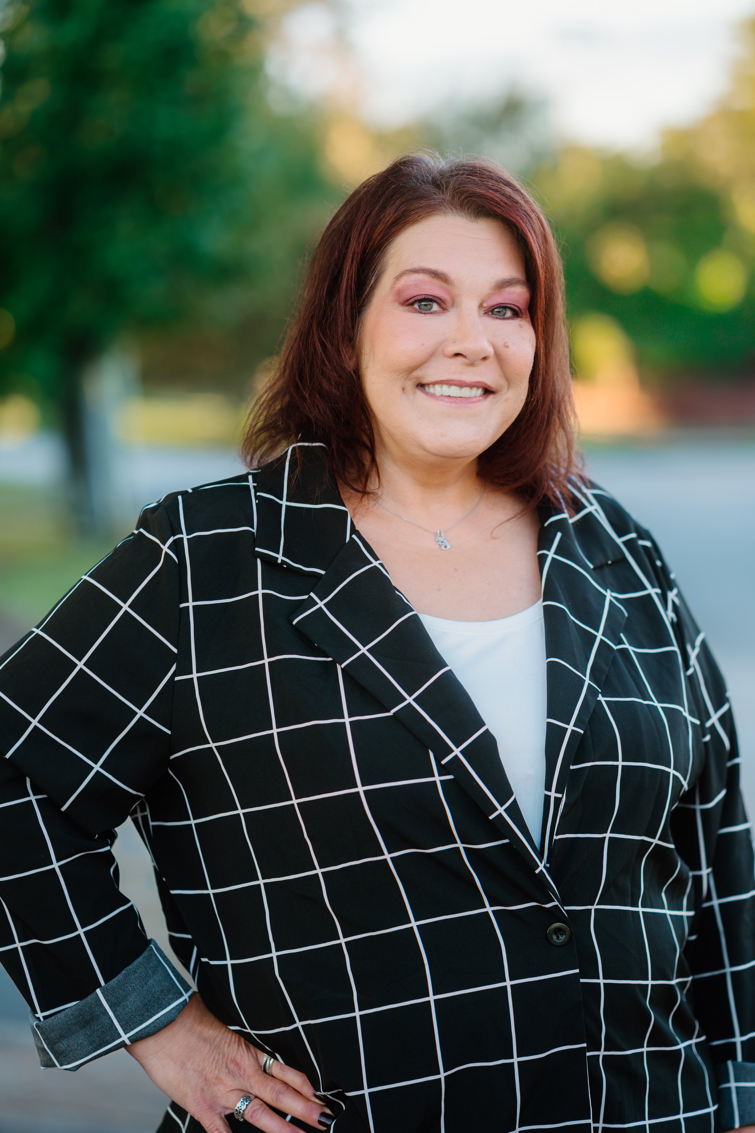A woman with shoulder-length brown hair outdoors, smiling, wearing a black blazer with white grid pattern over a white top, with trees and a street in the background.