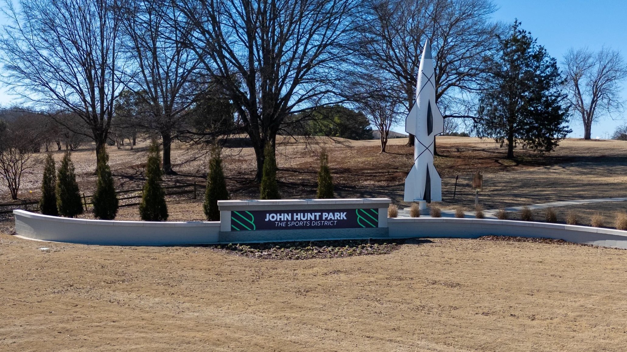 A landscaped park with a curving white wall, small trees, and a modern abstract sculpture standing on a circular base, with a grass area and a pathway leading to the distance, surrounded by trees and a street.