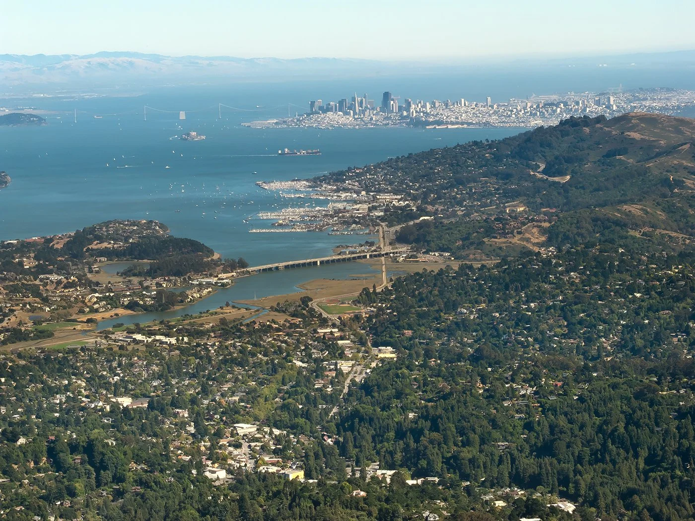 Aerial view of San Francisco Bay with the city skyline and Golden Gate Bridge in the distance, surrounded by hills and residential neighborhoods.