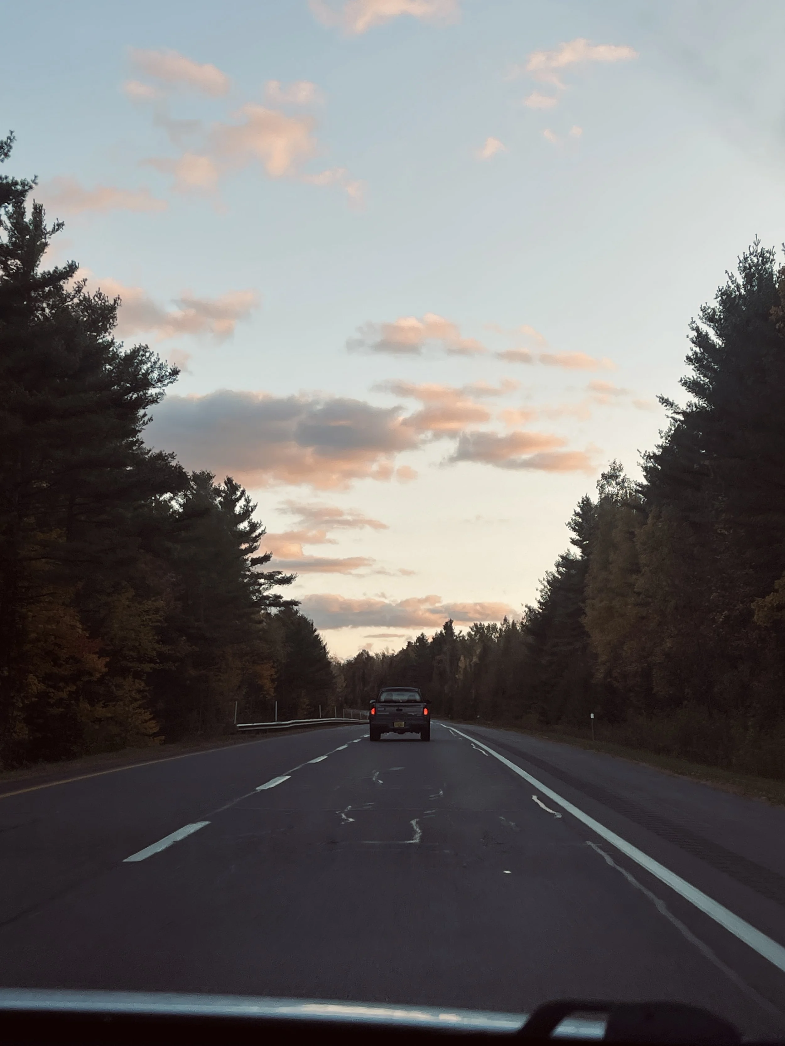 A black pickup truck driving down a two-lane highway lined with trees on both sides during sunset with a partly cloudy sky.