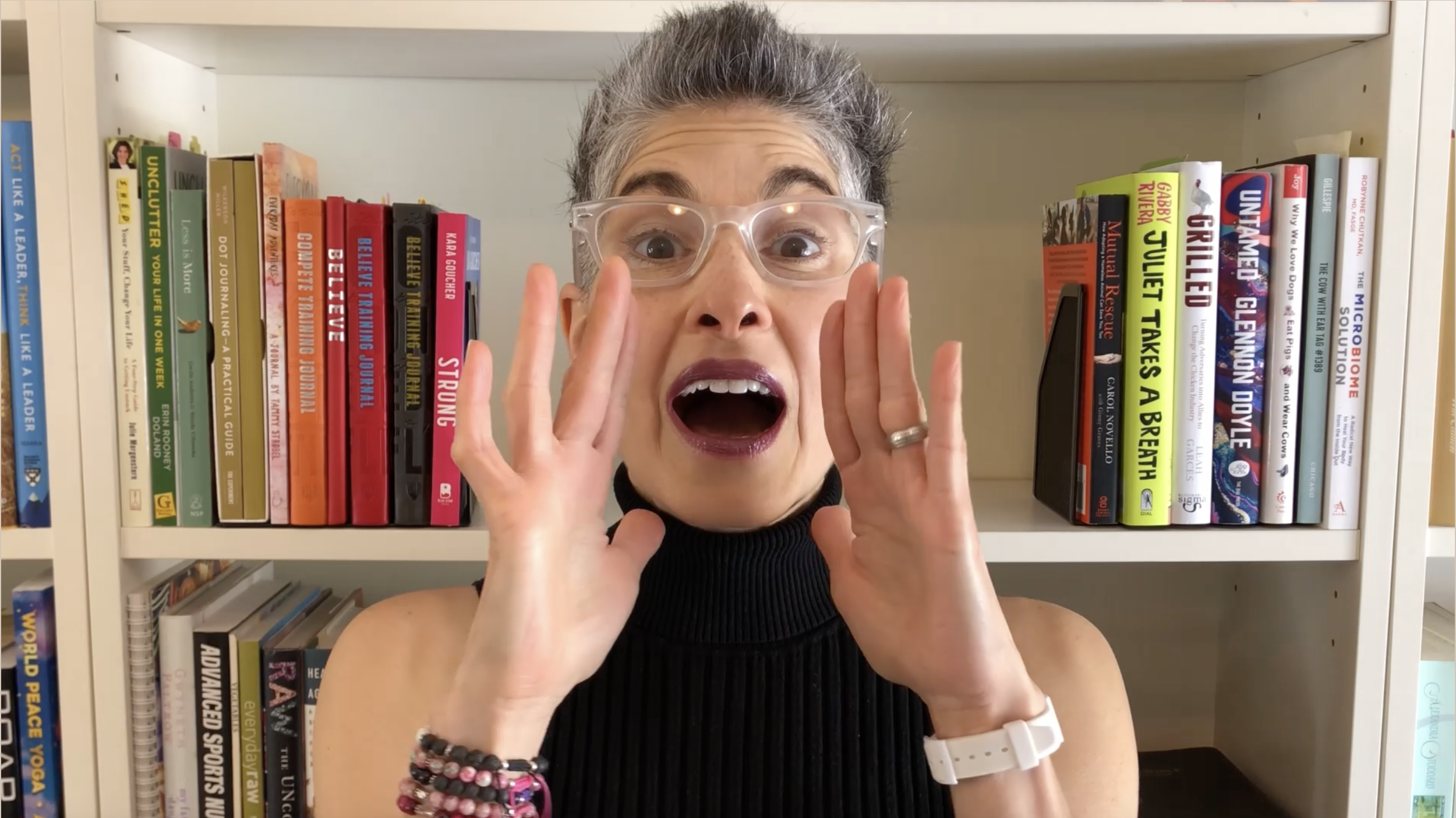 A woman with short gray hair and glasses holding her hands to her face, wide-eyed and open-mouthed, standing in front of a bookshelf filled with colorful books.