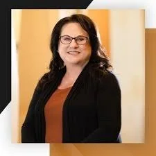 A woman with glasses and long dark hair smiling, wearing a black blazer and an orange top, standing indoors in front of a light-colored wall.