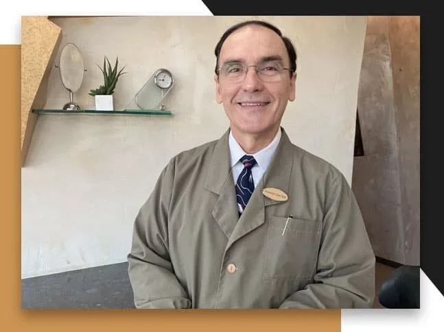 A smiling man in a taupe blazer, white shirt, and patterned tie standing in a modern interior space with a beige wall, a glass shelf with a small plant, a mirror, and a clock behind him.