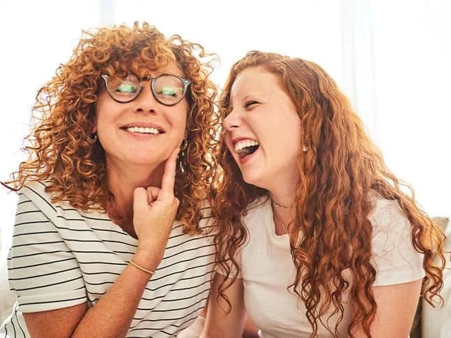 Two women with curly red hair laughing and smiling, one wearing glasses and a striped shirt, the other in a white shirt, indoors with bright lighting.