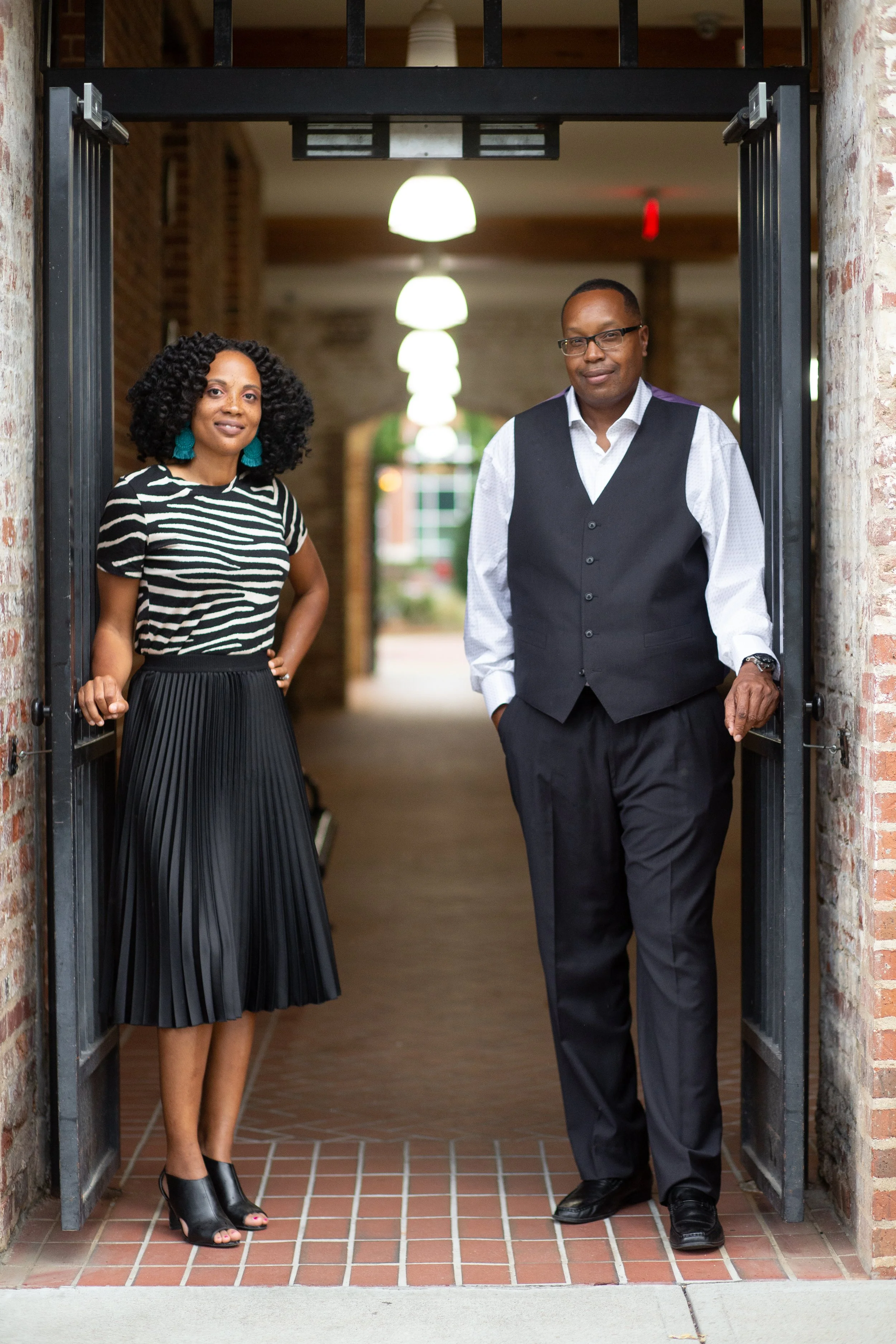 A woman and a man standing in a doorway inside a building, with the woman wearing a black and white striped top and black pleated skirt, and the man dressed in a white shirt with a black vest and black pants.
