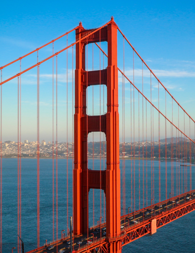 Aerial view of the Golden Gate Bridge in San Francisco, California, with the bridge's iconic red towers, suspension cables, and traffic below, against a blue sky and water.