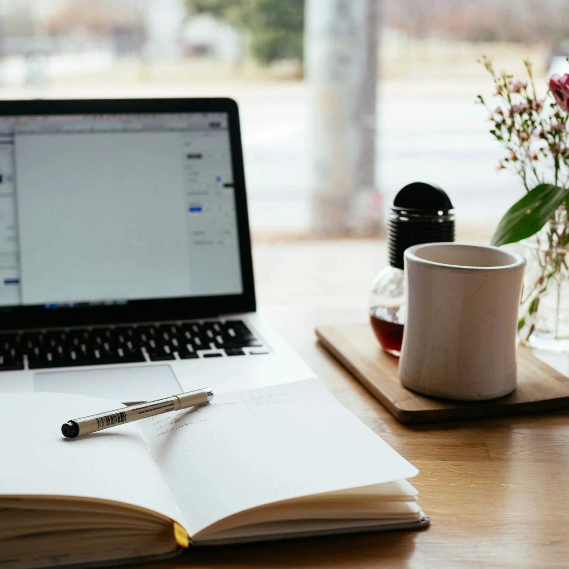A minimalist workspace with an open notebook and a pen on a wooden desk, a laptop displaying a blurred webpage, a white ceramic mug, a small glass jar, a soy sauce bottle, and a vase with pink flowers near a window.