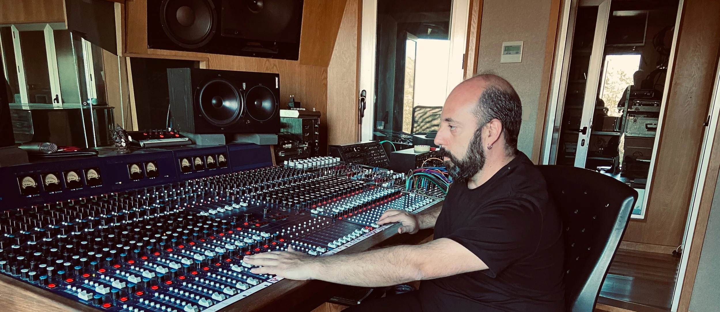 A man with a beard and bald head sitting in front of a large sound mixing console in a recording studio, surrounded by studio equipment, with wood-paneled walls and a glass door in the background.