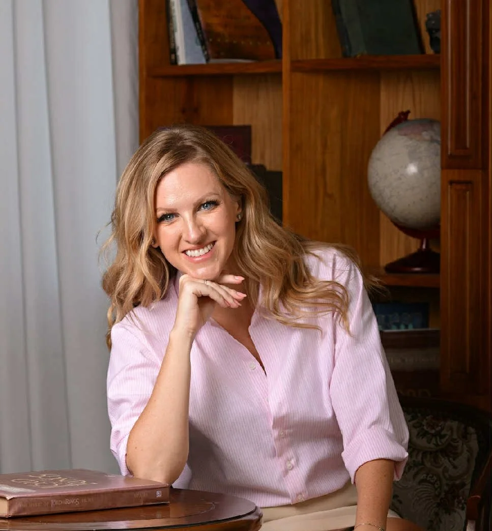 A woman with long, wavy blonde hair sitting at a wooden table in a room with a bookshelf behind her, holding a book and smiling at the camera.