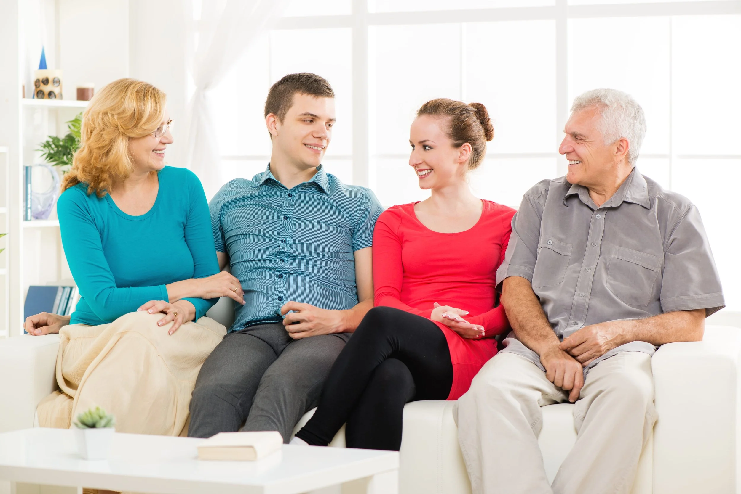 Five people sit on a white couch in a bright living room, happily talking and smiling at each other.