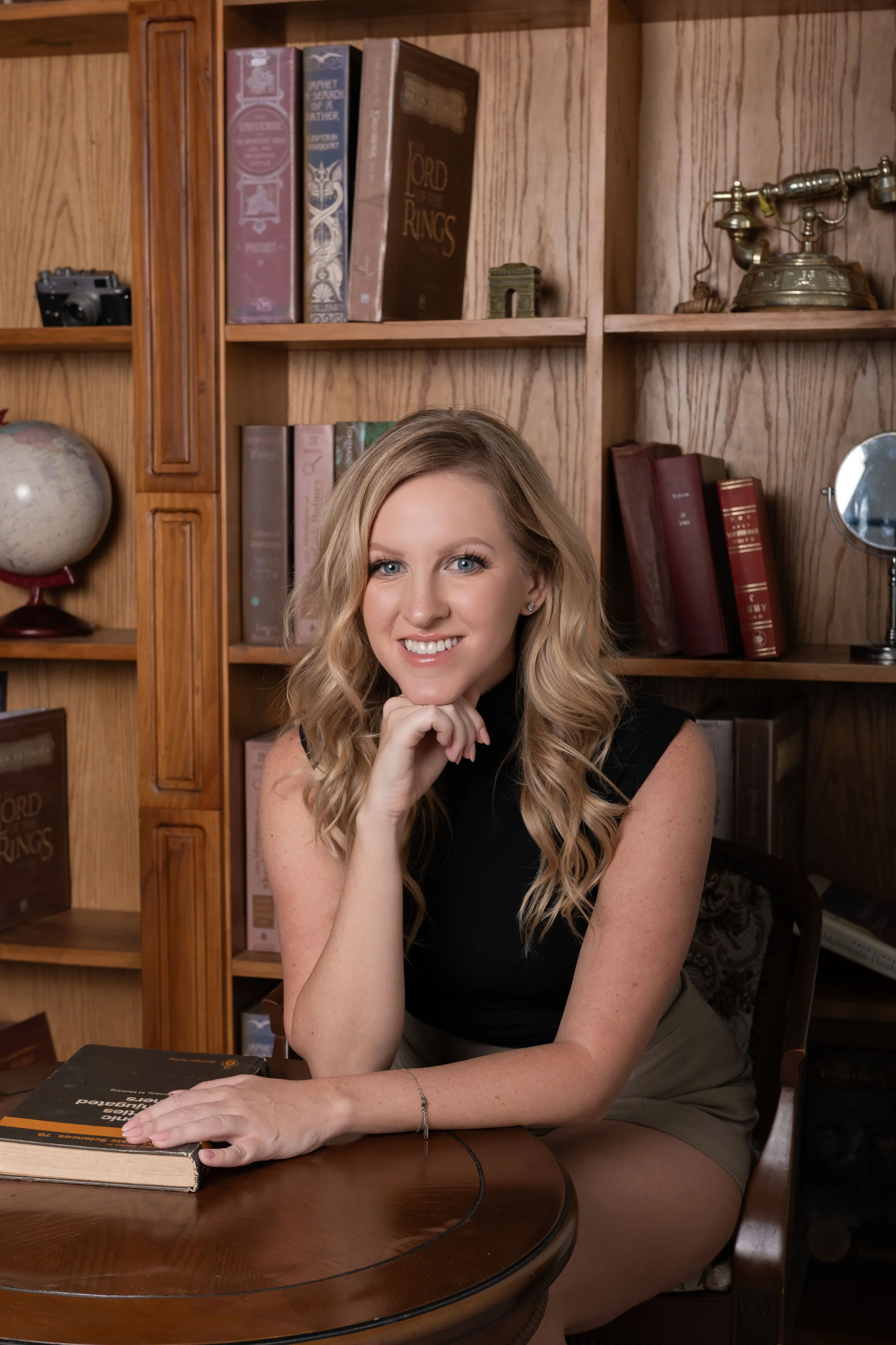 A woman with blonde wavy hair, wearing a black sleeveless top, sits at a wooden table with her left hand resting on a book, against a background of wooden bookshelves filled with books, a globe, an antique telephone, and a small mirror.