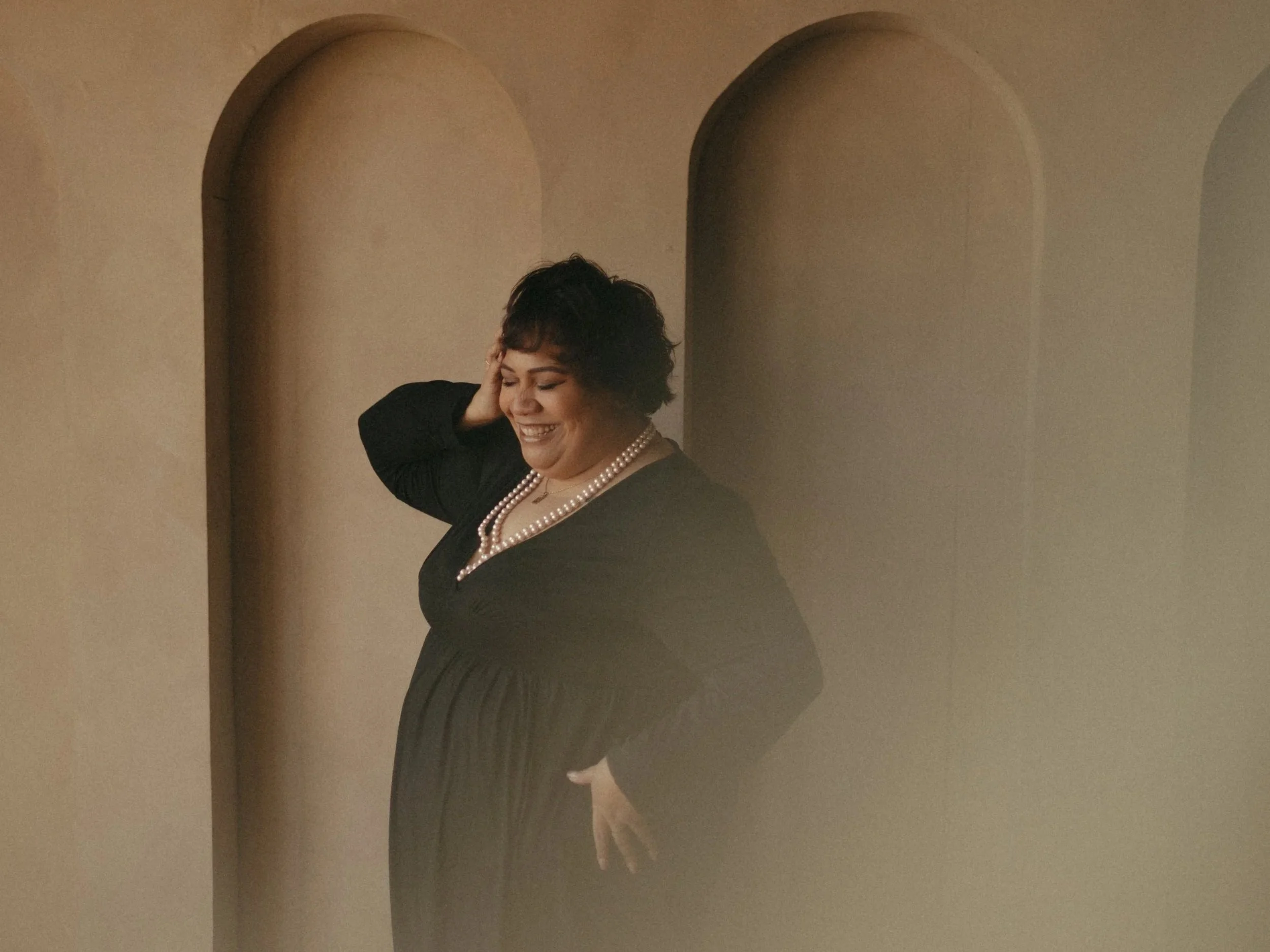 A woman with short dark hair, wearing a black dress and a pearl necklace, smiling and touching her neck, standing in front of arched wall niches.