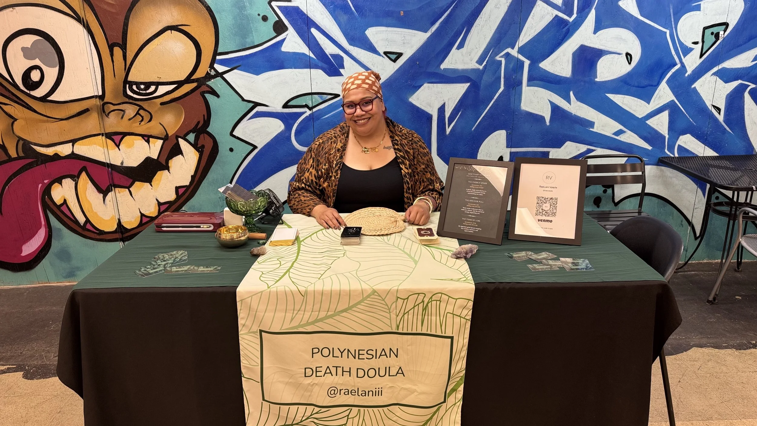 Woman sitting at a table with Polynesian Death Doula sign, tarot cards, crystals, and spiritual items, in front of a colorful graffiti mural.