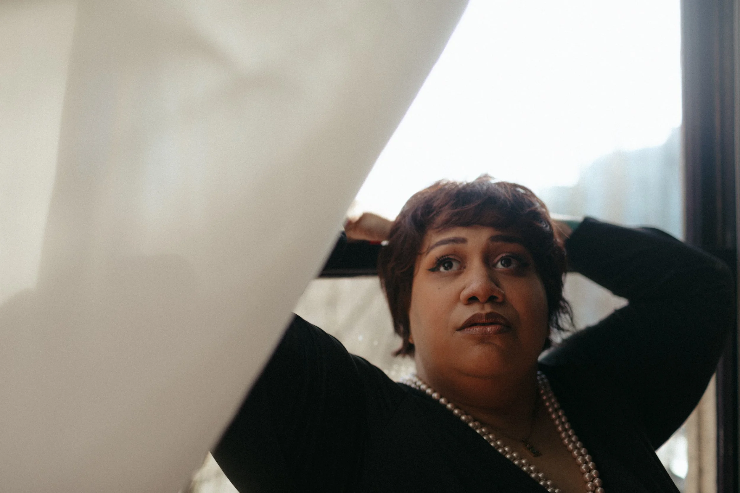 photo of rae with short hair and pearl necklace resting her hands behind her head, sitting by a window.