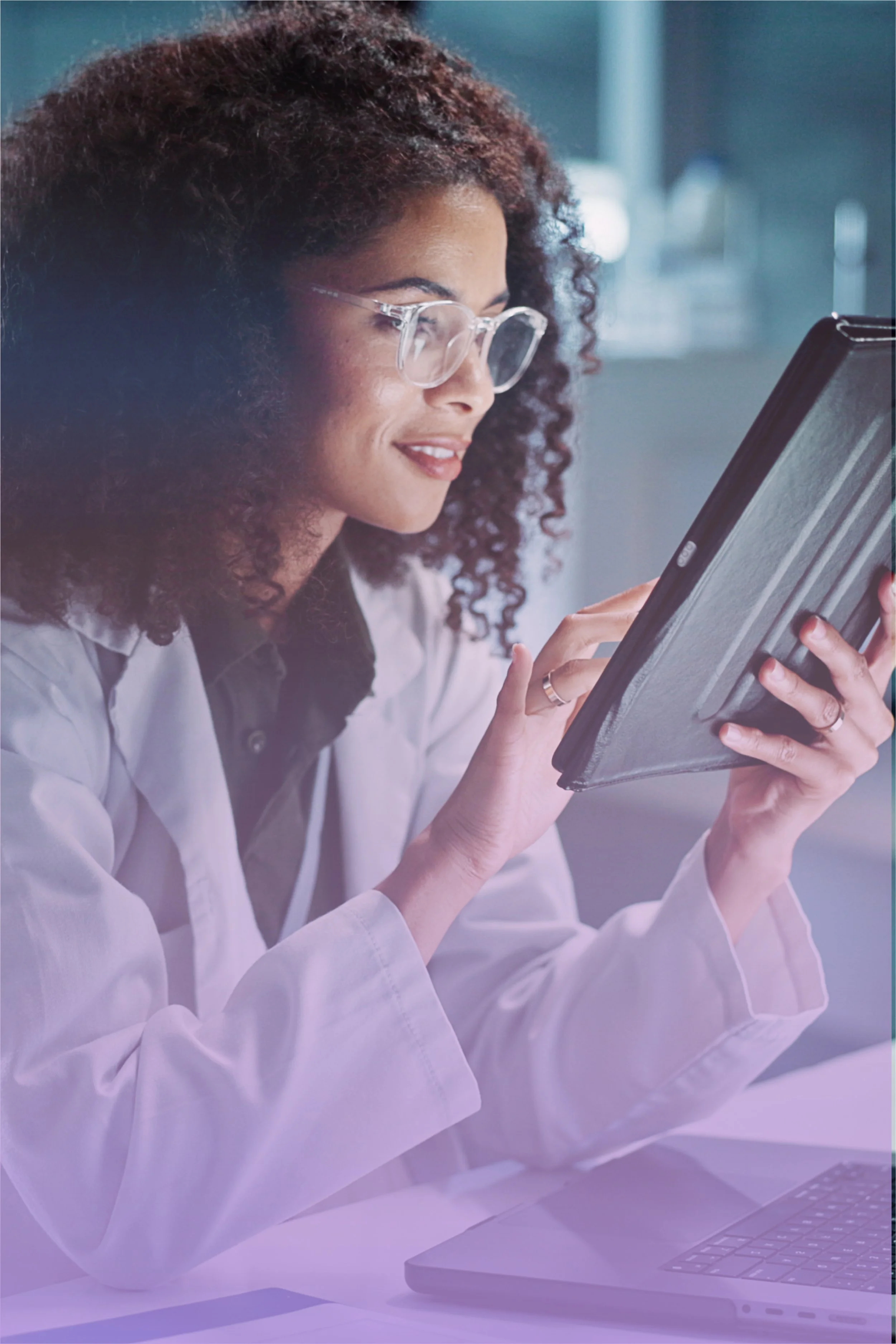 A woman with curly hair and glasses wearing a white lab coat, looking at a tablet device.