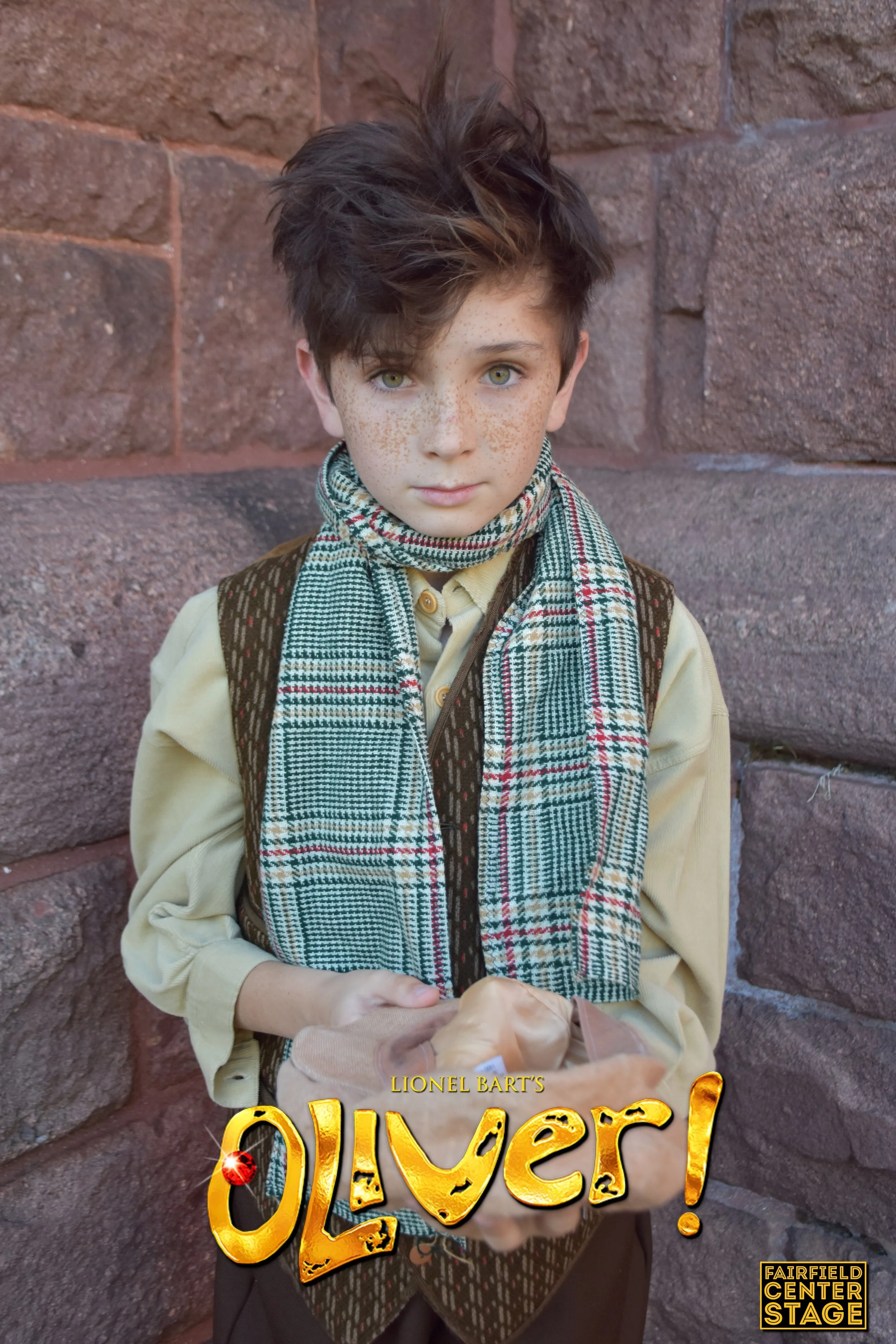A young boy with messy brown hair, wearing a plaid scarf, yellow shirt, and brown vest, standing against a stone wall, holding a hat with both hands, with the text 'Oliver!' at the bottom and a small yellow logo that says 'Fairfield Center Stage'.