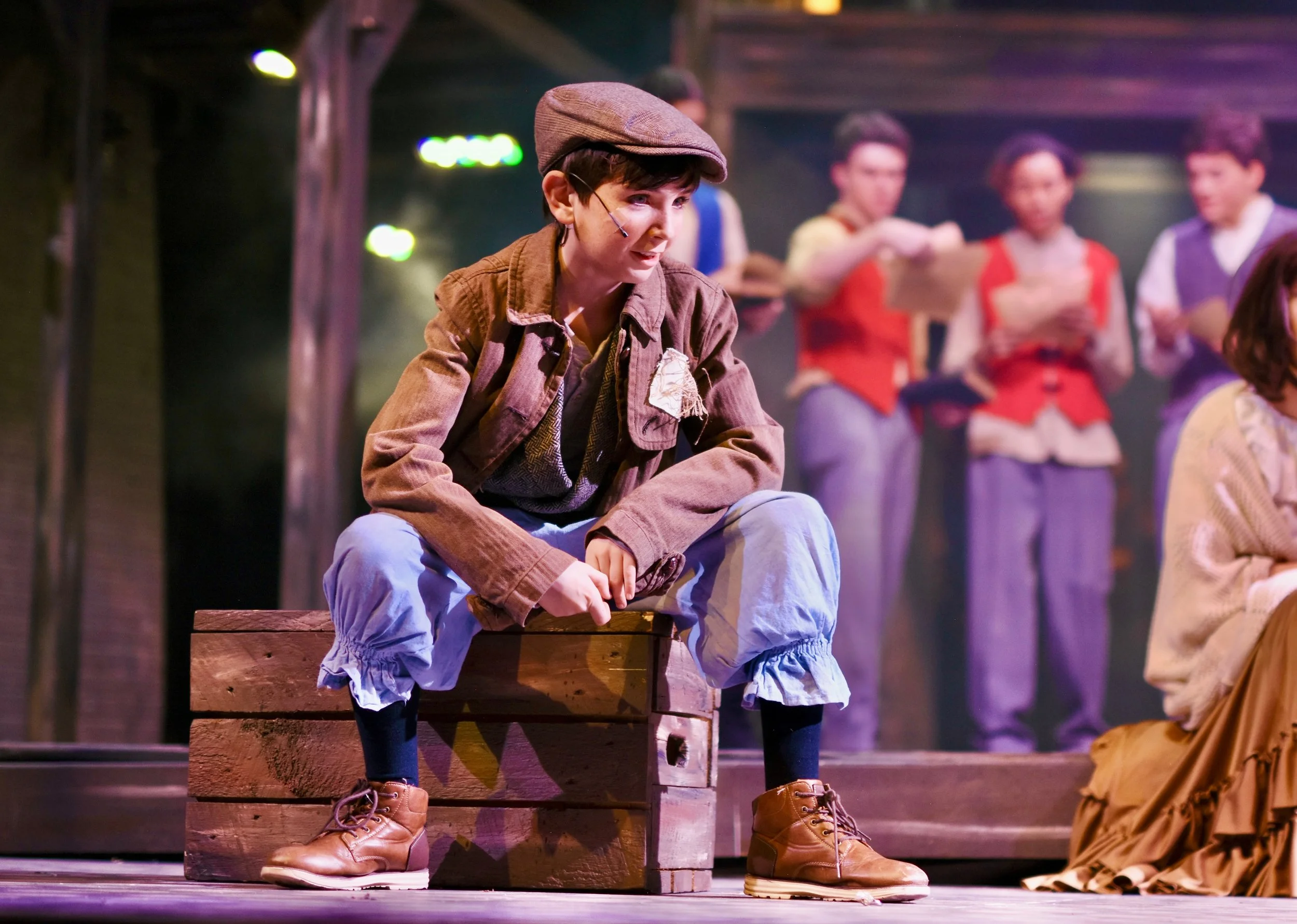 A person dressed in old-fashioned clothing, including a cap and brown jacket, sitting on a wooden crate on stage, with a group of people in similar period costumes in the background.