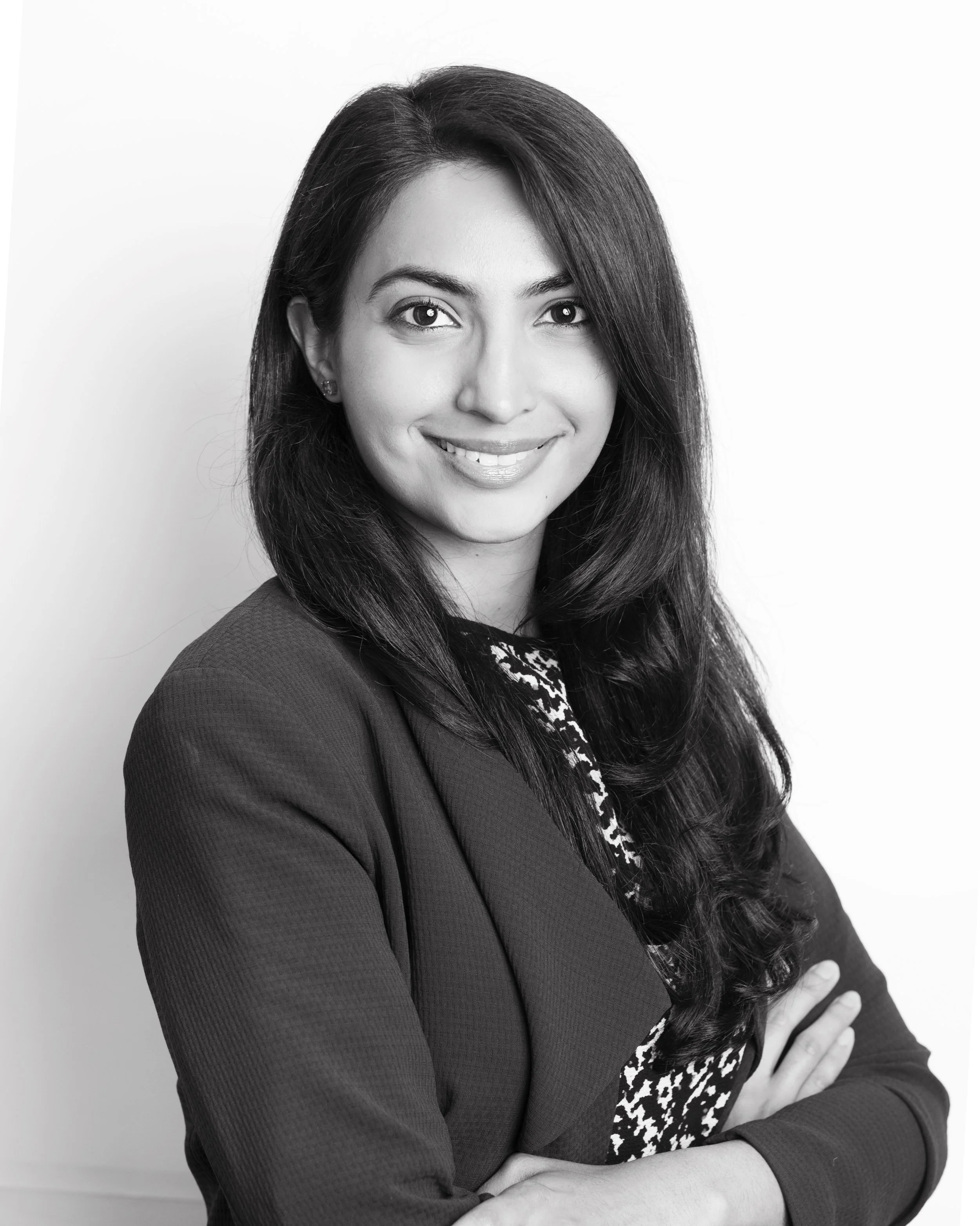 Black and white professional portrait of a woman with long dark hair, smiling, wearing a blazer and patterned top, standing with arms crossed against a plain background.
