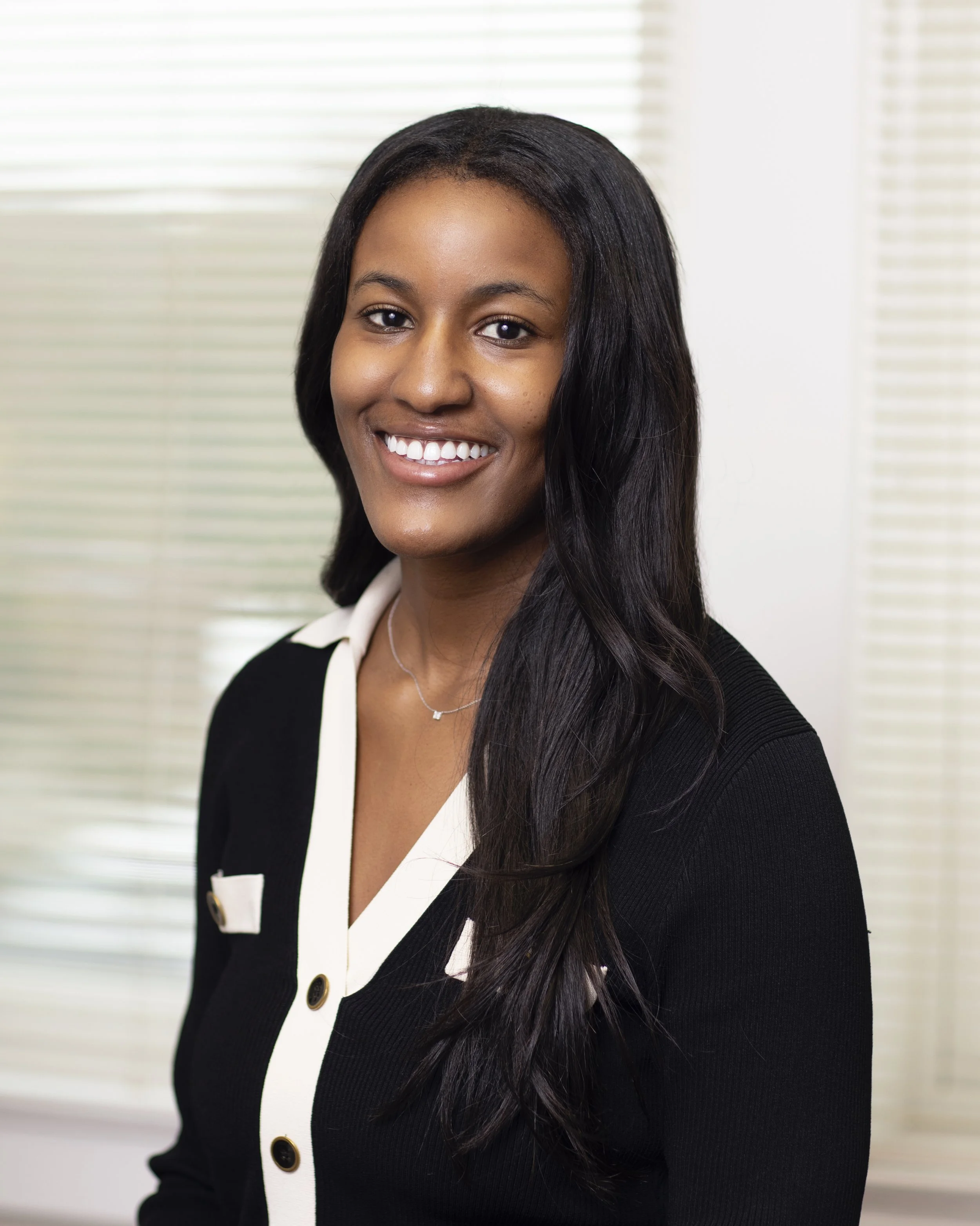 A young woman with long dark hair, smiling, wearing a black and white blazer, standing indoors in front of blinds.