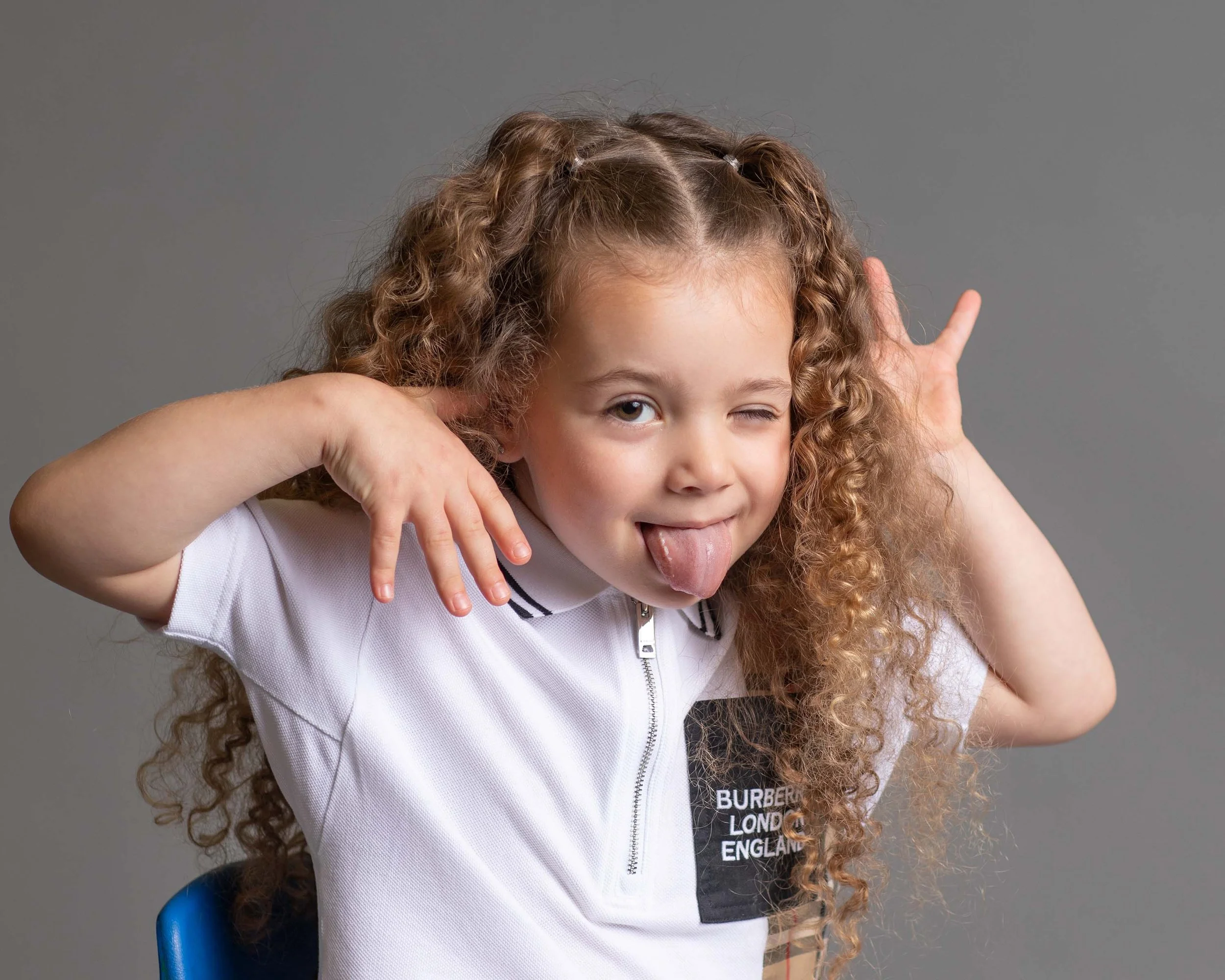 A young girl with curly hair making a playful face, sticking out her tongue and winking, while holding her hands near her head with fingers spread.