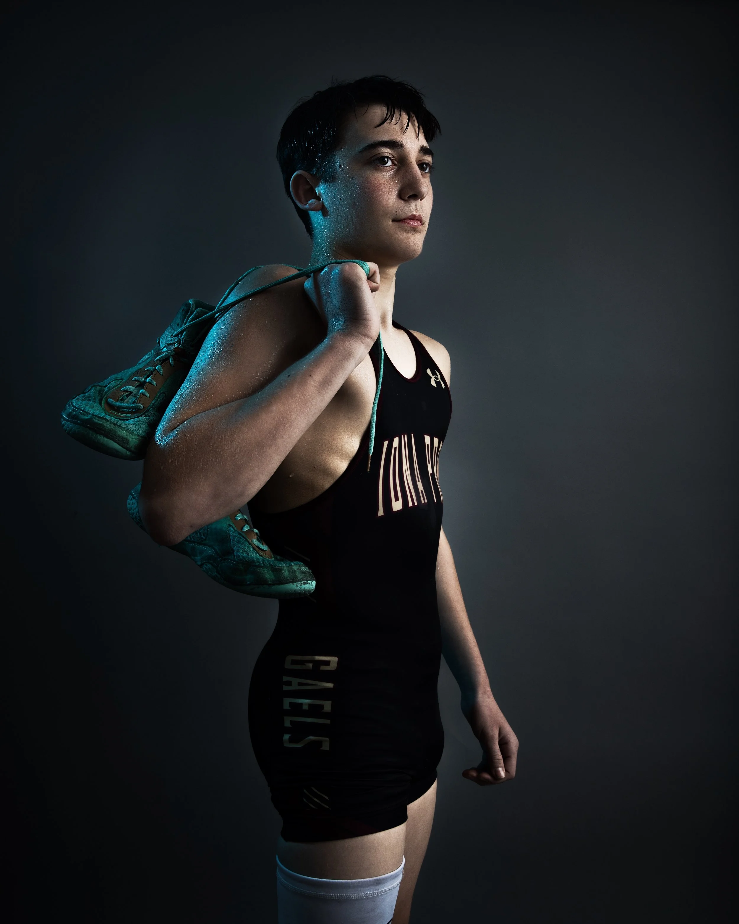 Young female athlete in athletic gear with a shoe over her shoulder, standing against a dark background.