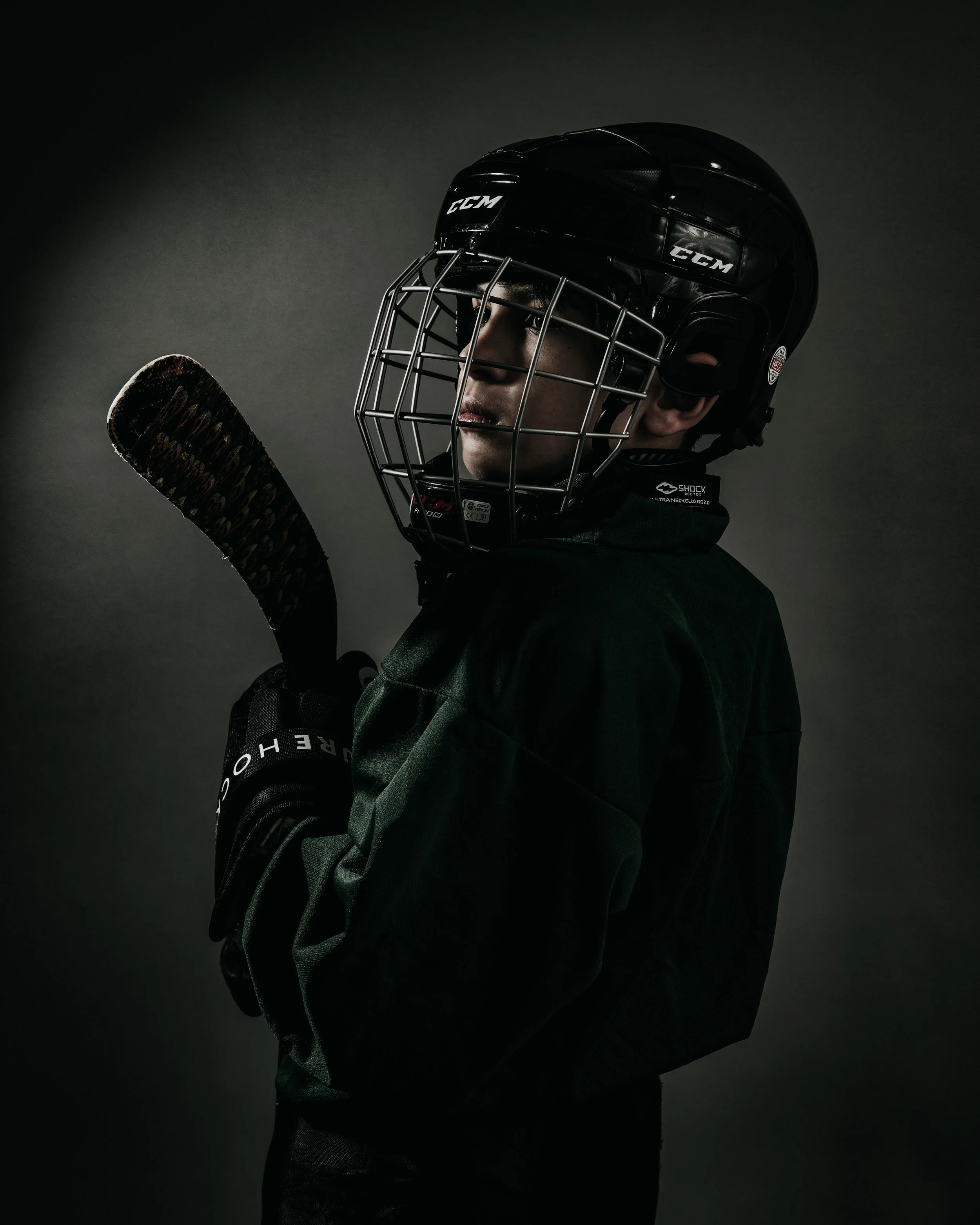 A hockey player in a black helmet with a face cage, dark green jersey, and holding a hockey stick, standing in a dark background.