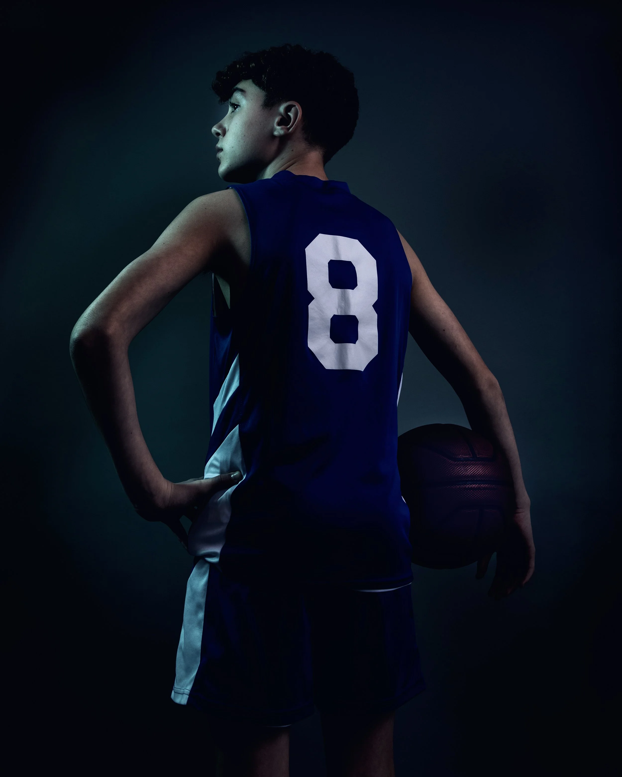 A young male basketball player with curly dark hair, wearing a blue jersey with the number 8, holding a basketball behind his right thigh, standing in profile against a dark background.