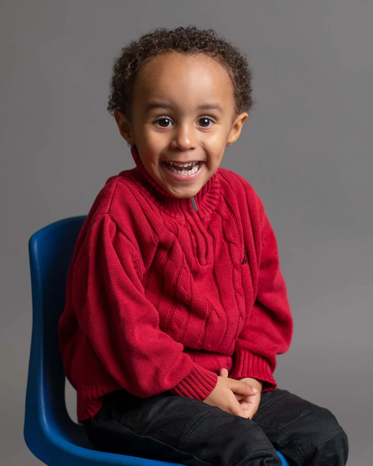 Young boy with curly hair smiling and wearing a red sweater, sitting on a blue chair against a gray background.