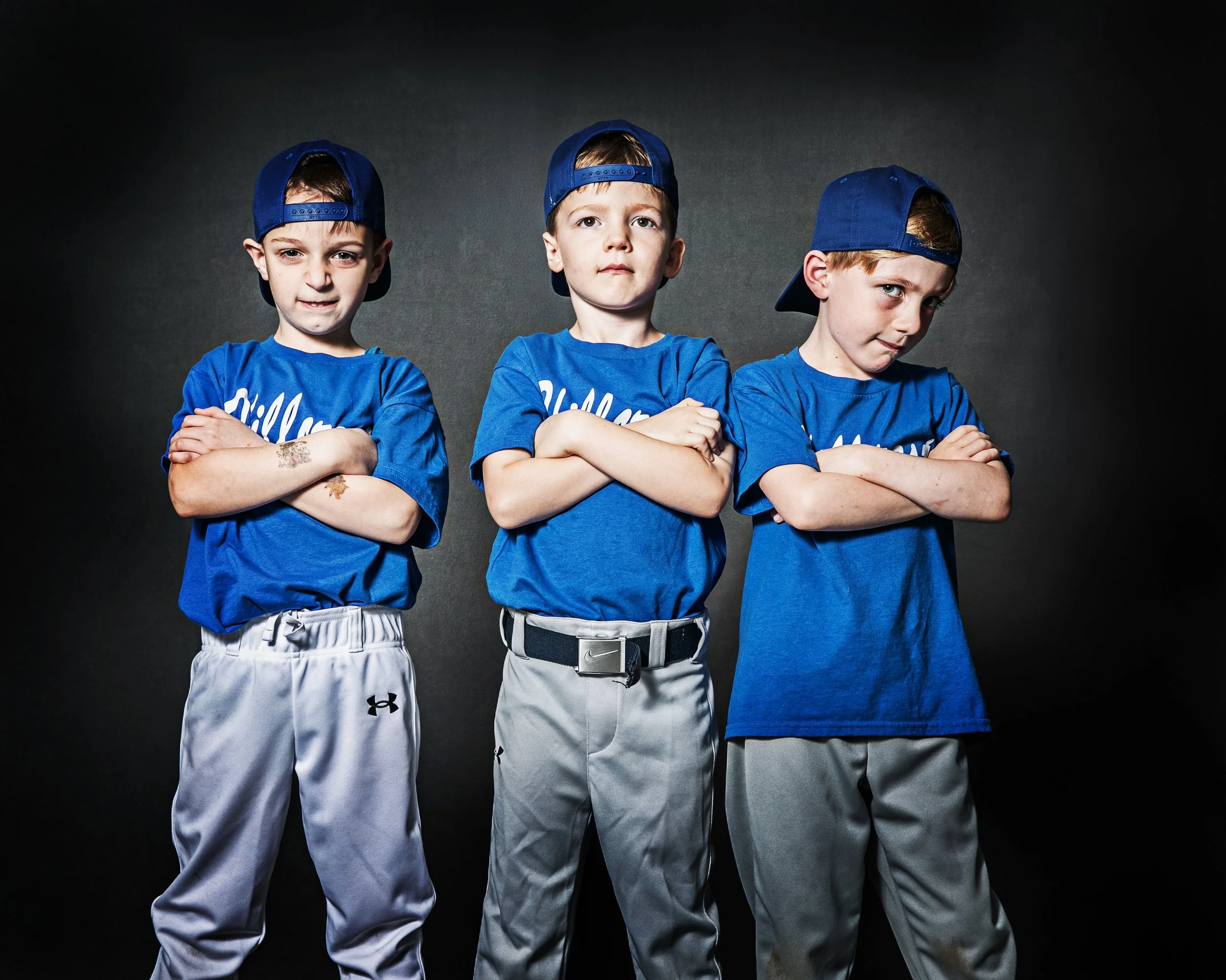 Three young boys wearing blue baseball jerseys and gray pants, standing with arms crossed, against a dark background.