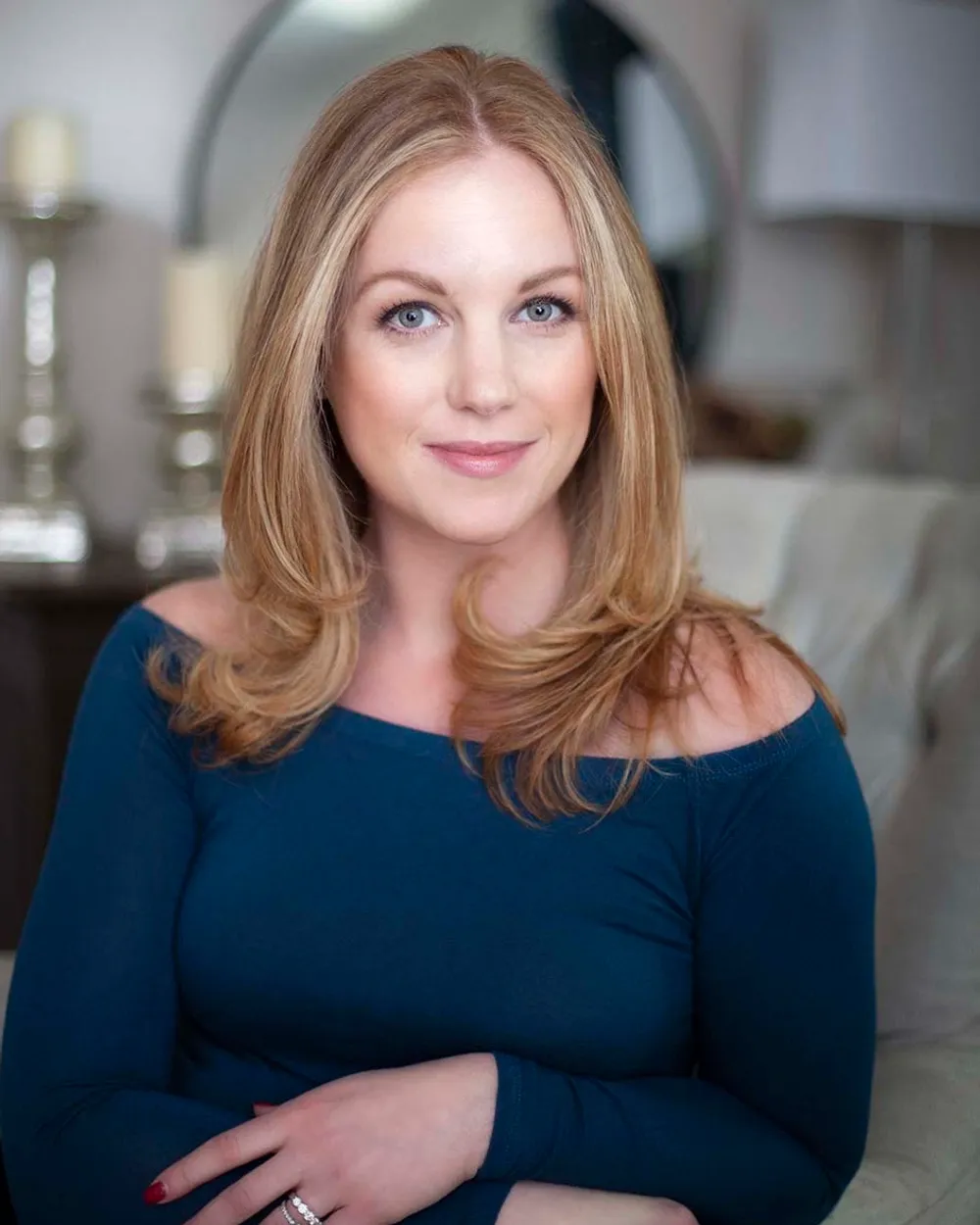 A woman with shoulder-length blonde hair and blue eyes, wearing a dark blue off-shoulder top, sitting on a beige sofa in a living room with candles and decorative items in the background.