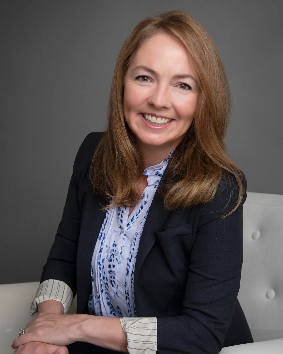 A woman with long red hair, smiling, wearing a dark blazer over a light blue patterned blouse, sitting on a white chair with a gray background.