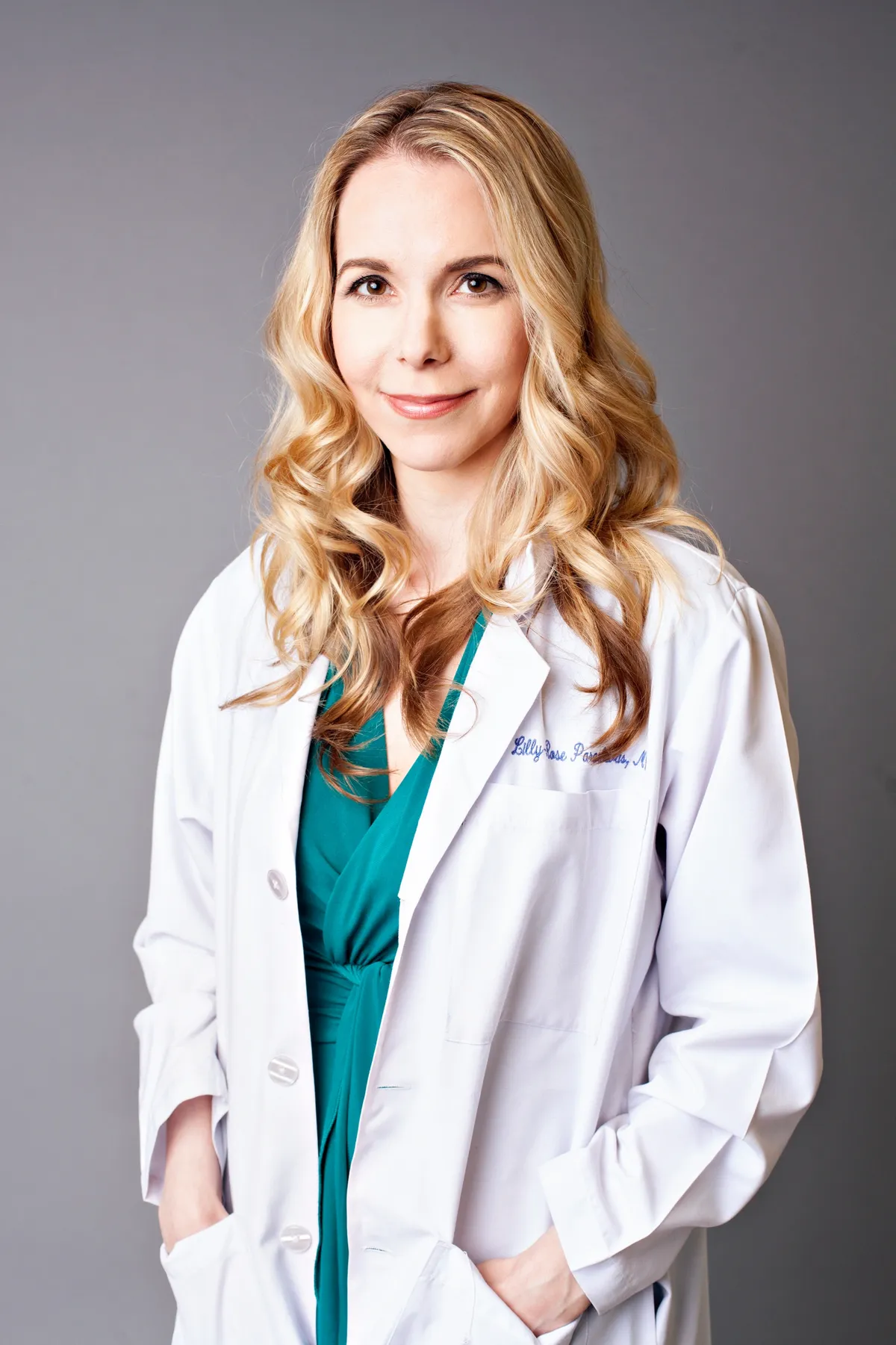 Portrait of a female doctor with blonde curly hair, wearing a white lab coat over a teal top, standing against a gray background.