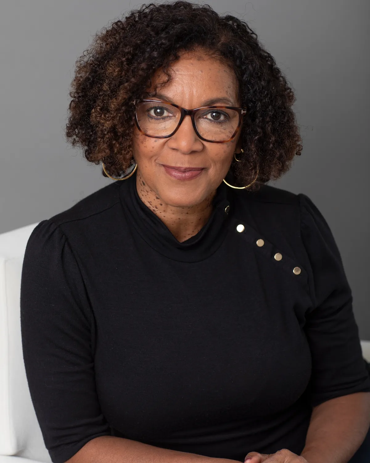 A woman with curly hair, wearing glasses and a black top with gold accents, sitting against a gray background.