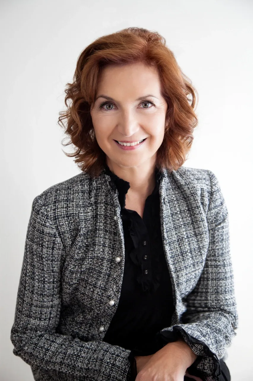 Portrait of a woman with wavy red hair, wearing a gray tweed blazer and black top, smiling at the camera against a plain white background.