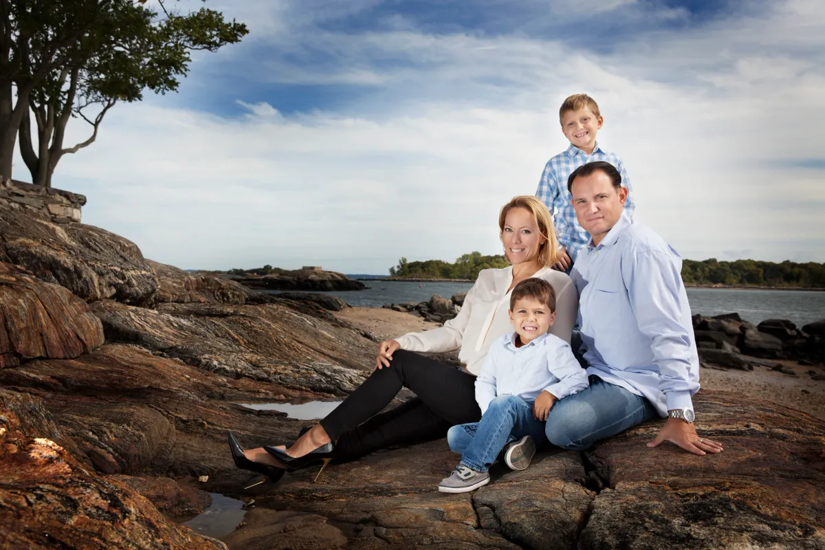 Family of four sitting on rocks by the water with a tree on the left and a cloudy sky.