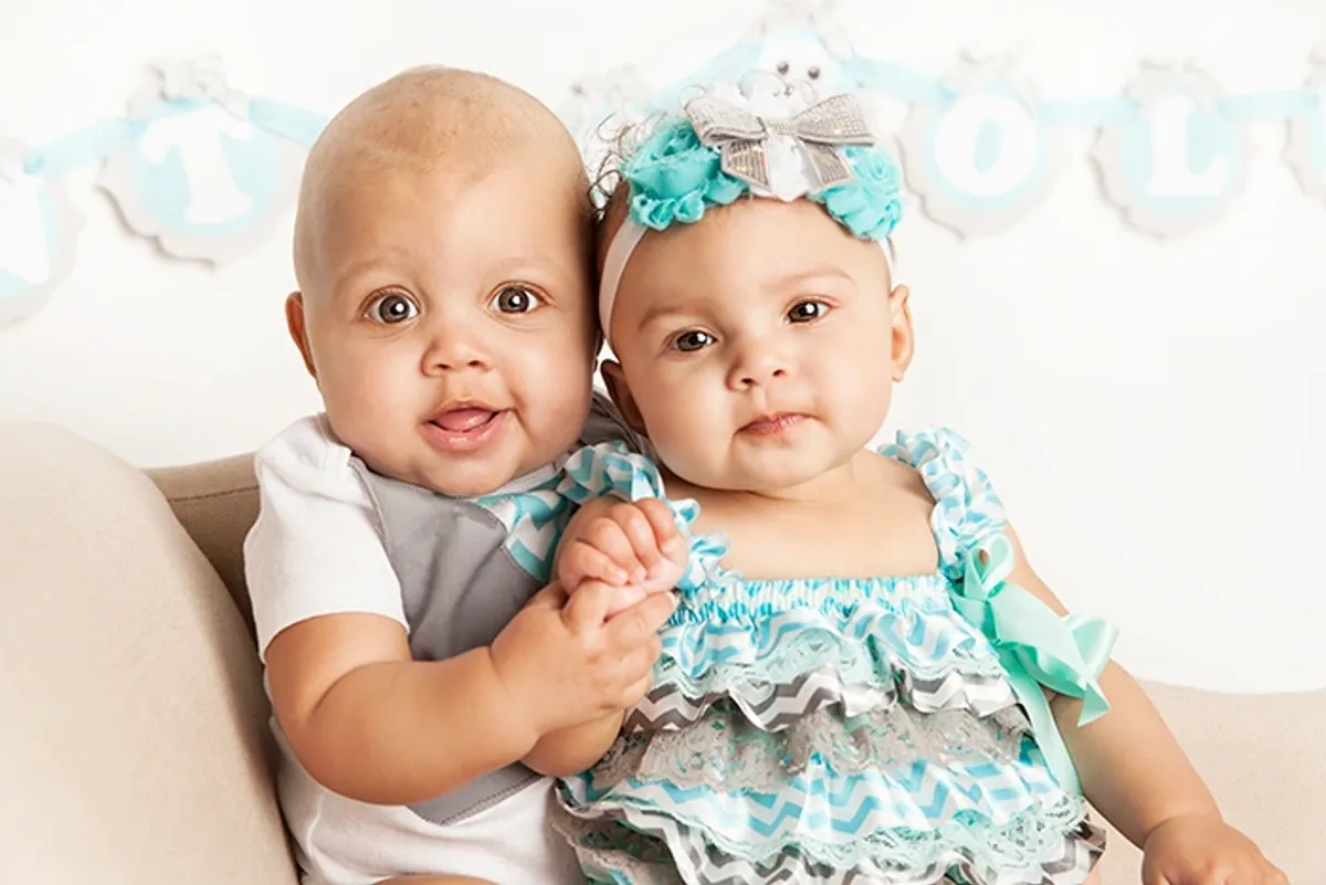 Two babies sitting close together on a beige couch, holding hands, with a blue and white birthday banner in the background. The baby on the left has a bald head, big eyes, and is sticking out her tongue. The baby on the right has curly hair, a large 