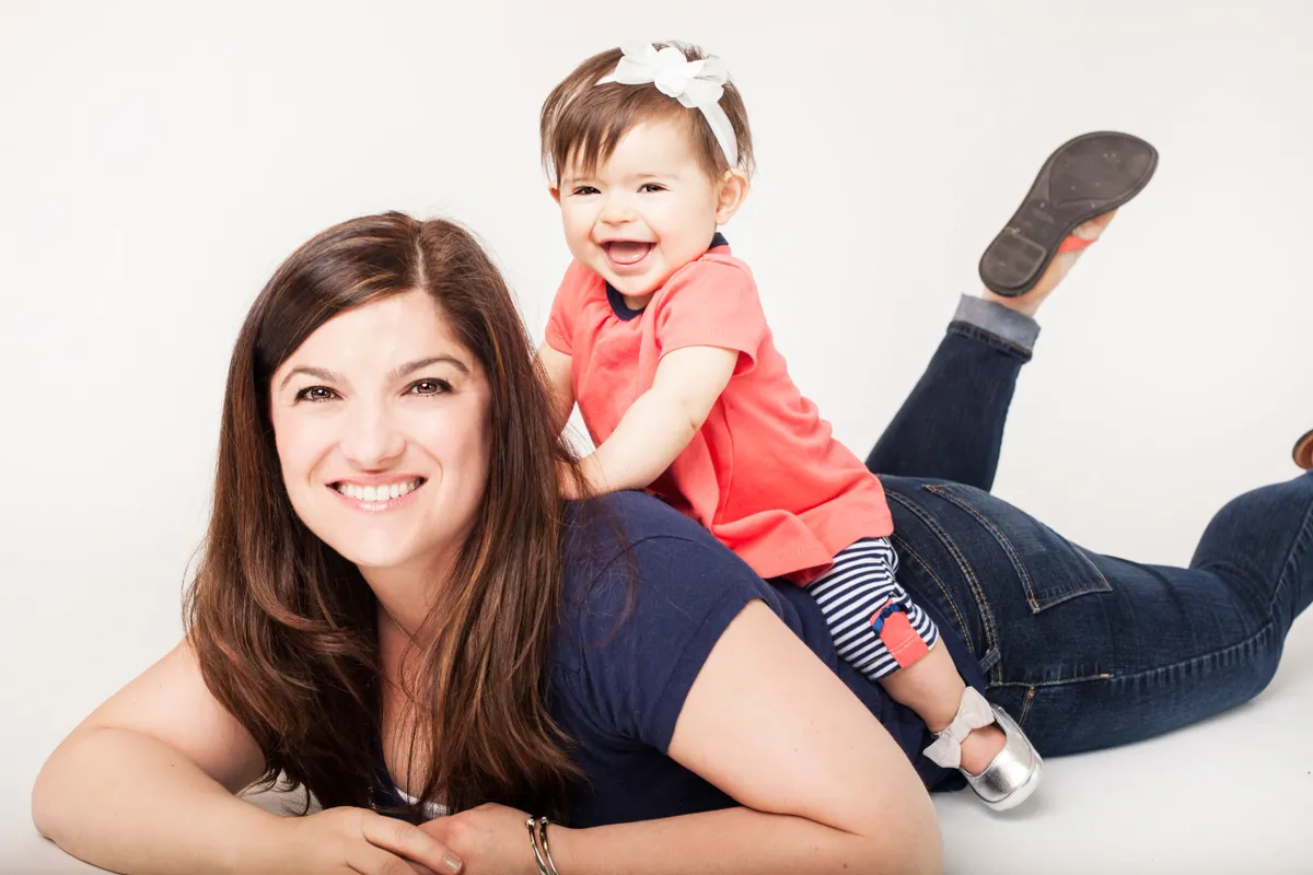 A woman lying on her stomach with a young girl on her back, both smiling and looking at the camera against a white background.
